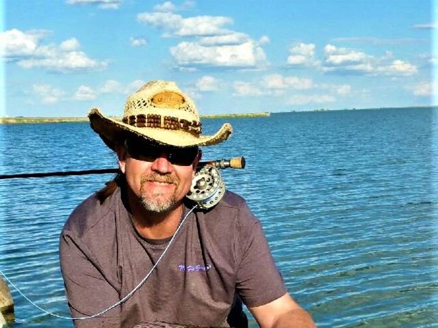 Man in cowboy hat and sunglasses fishing by a calm lake.