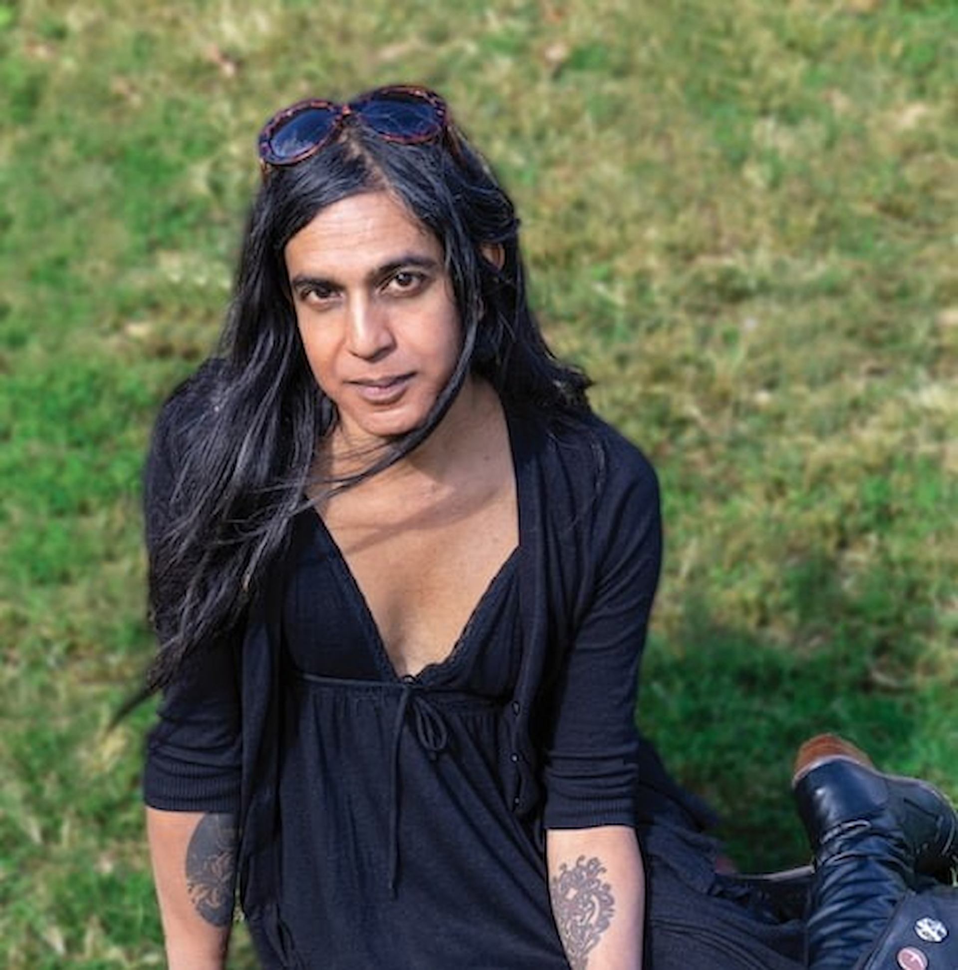 Bishakh Som seated on grass in black outfit with sunglasses and visible tattoos.