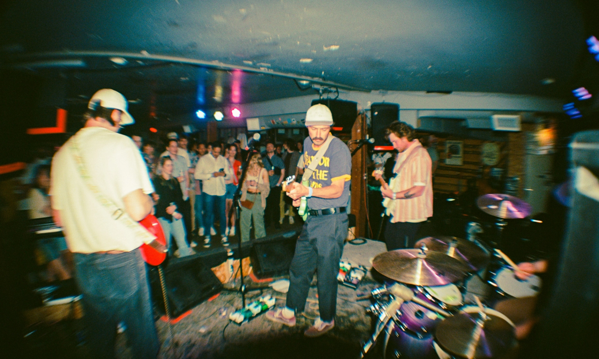 Indie rock band performs on small club stage as crowd watches near the musicians.
