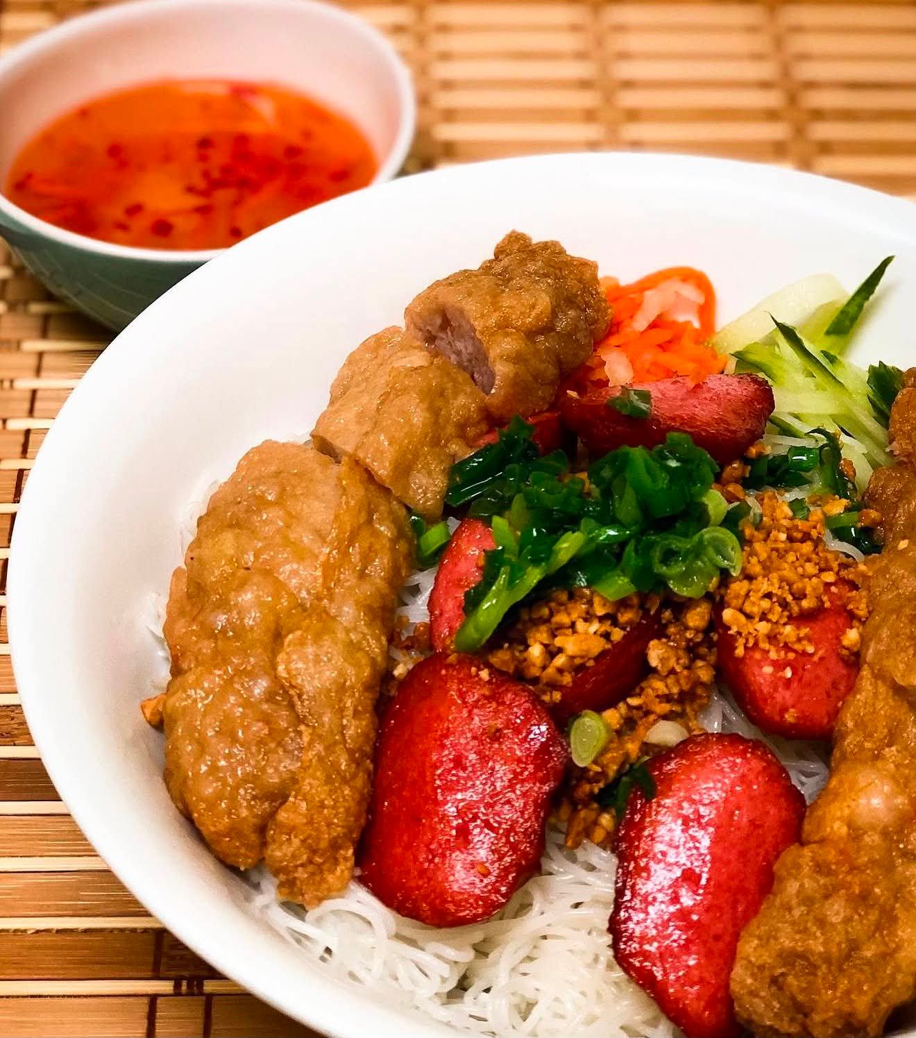 Bowl of vermicelli noodles with fried rolls, sausage slices, vegetables and dipping sauce.