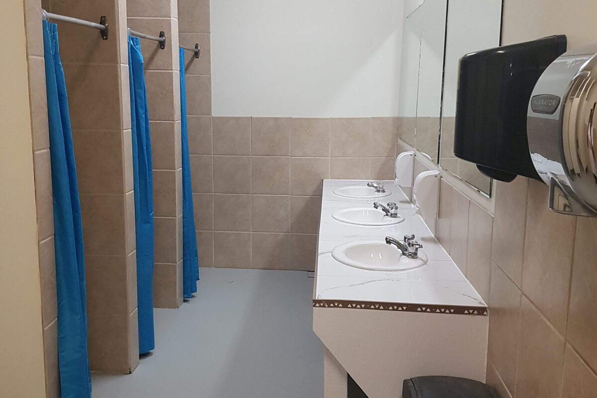 Washroom with three sinks, mirrors, tiled shower stalls, and blue curtains.