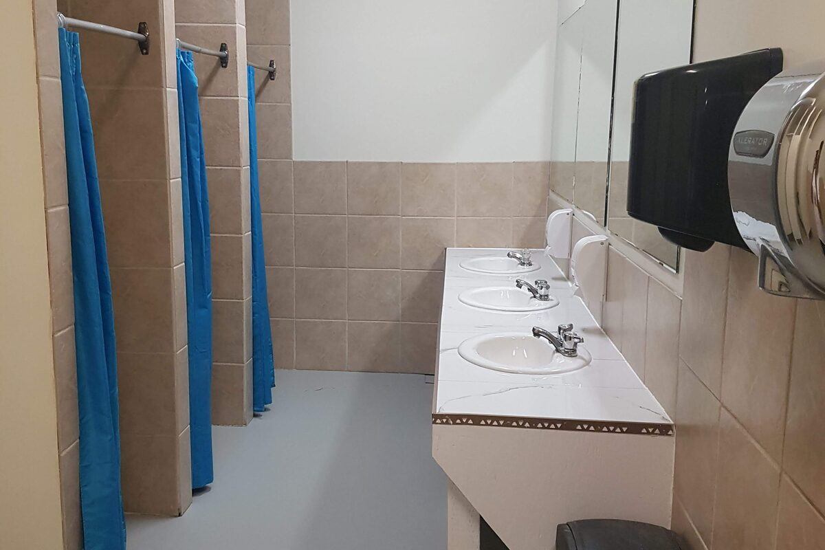 Washroom with three sinks, mirrors, tiled shower stalls, and blue curtains.