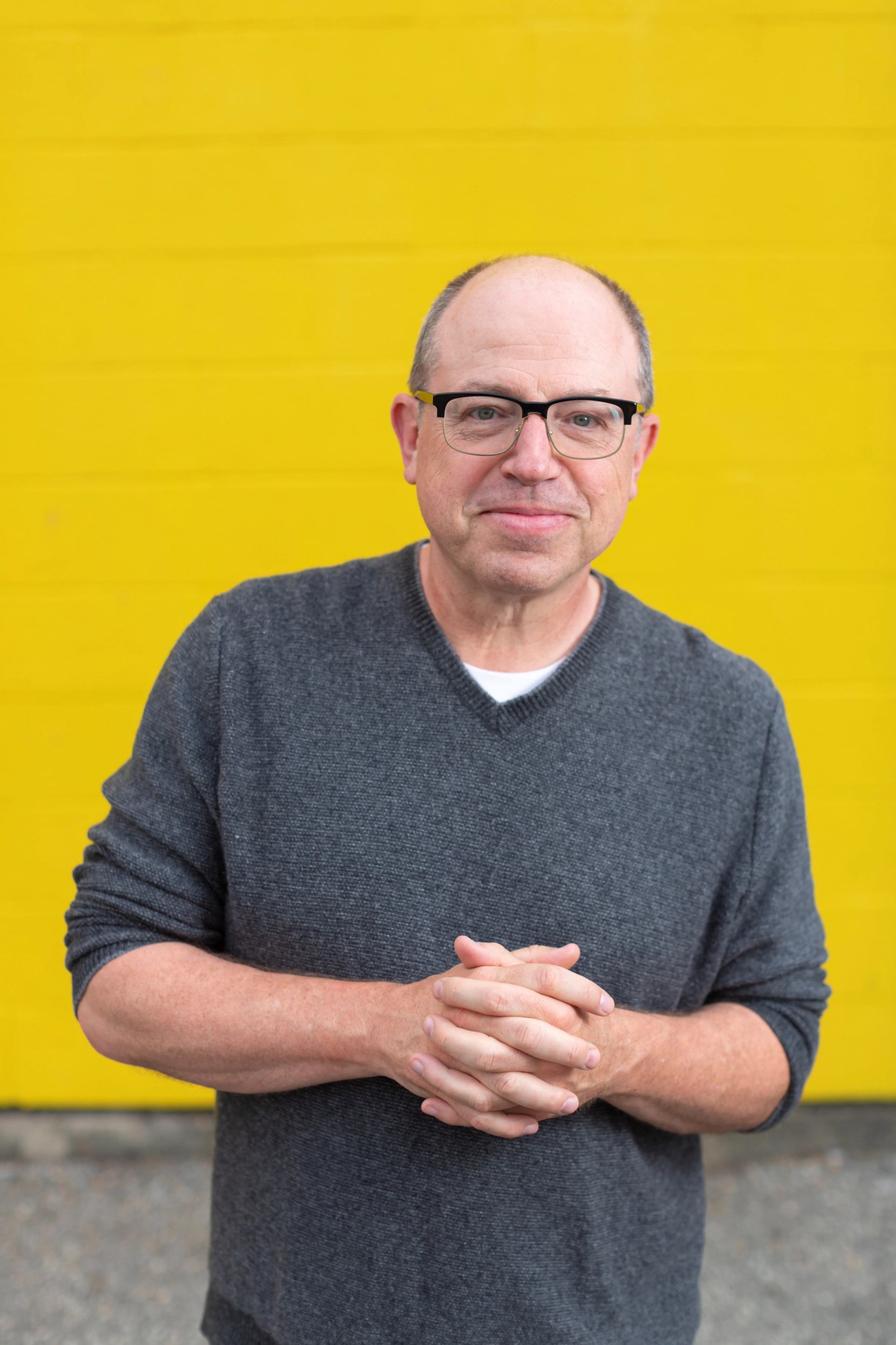 Person wearing glasses and grey sweater stands clasping hands before bright yellow wall.