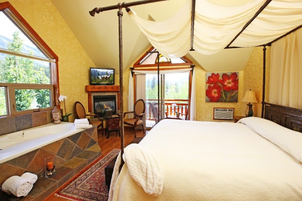 A cozy bedroom with a four-poster bed, fireplace, TV, a luxurious bathtub, and scenic mountain view.