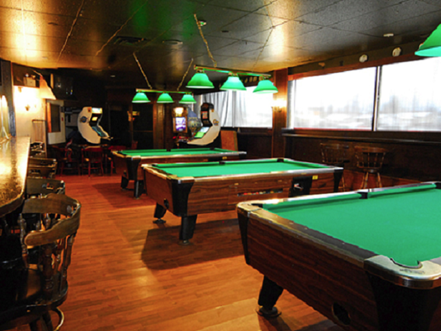 Dimly lit pool hall with three pool tables, green hanging lights, and arcade machines. Warm wood tones create a cozy, relaxed atmosphere.