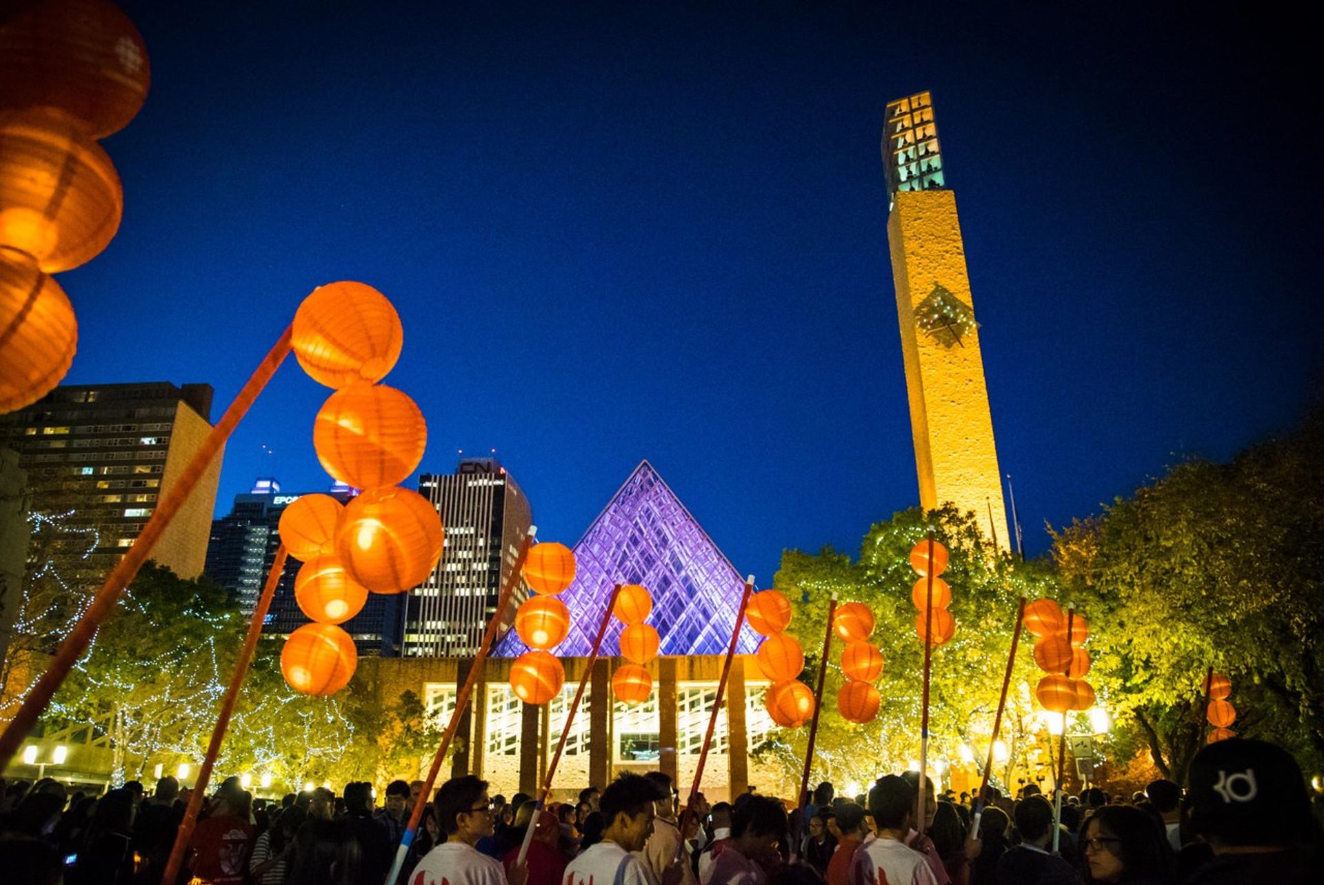 Crowd holding orange lanterns in city square with lit glass pyramid and tall tower.
