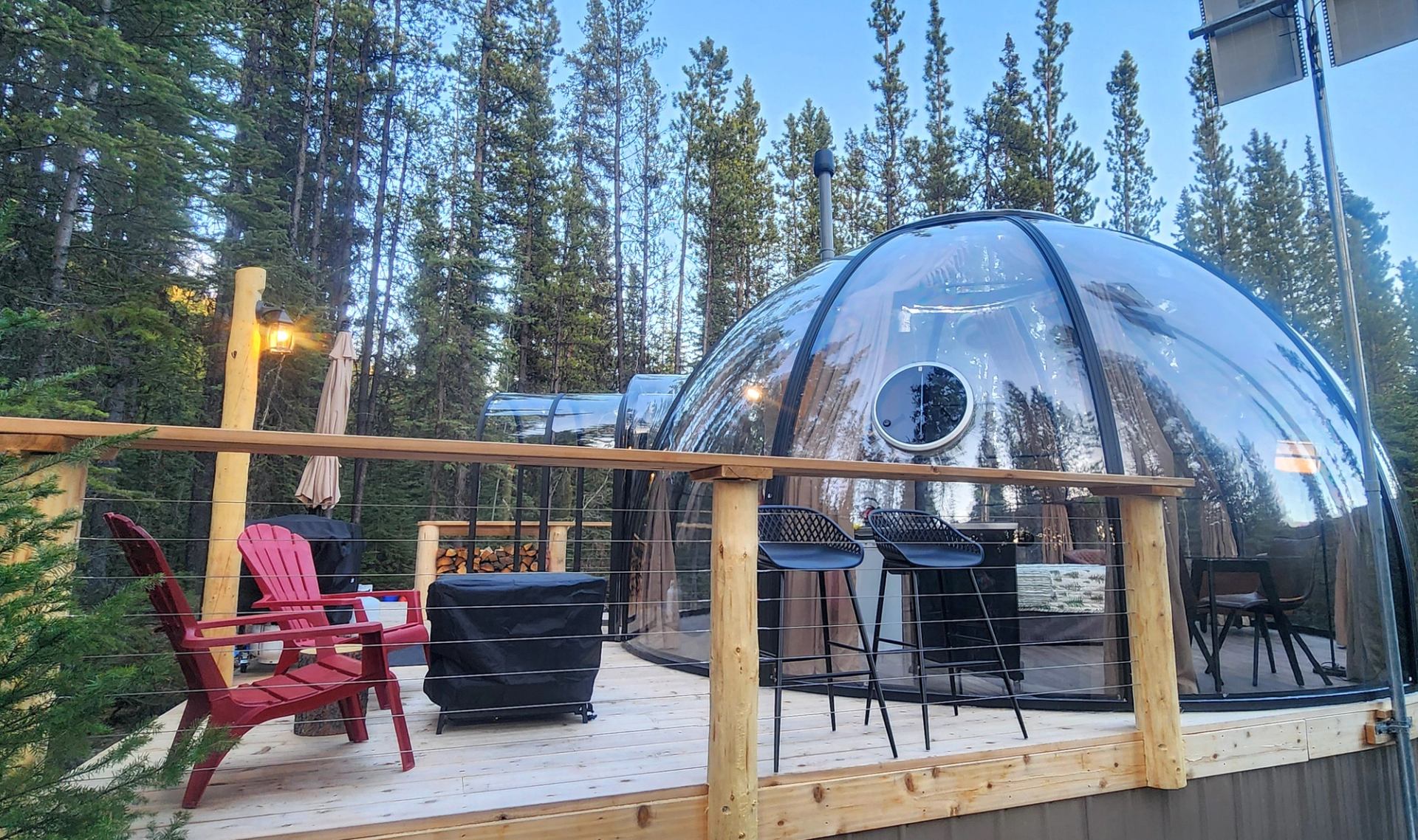 Transparent dome cabin with deck, red chairs and bar stools in forest setting.