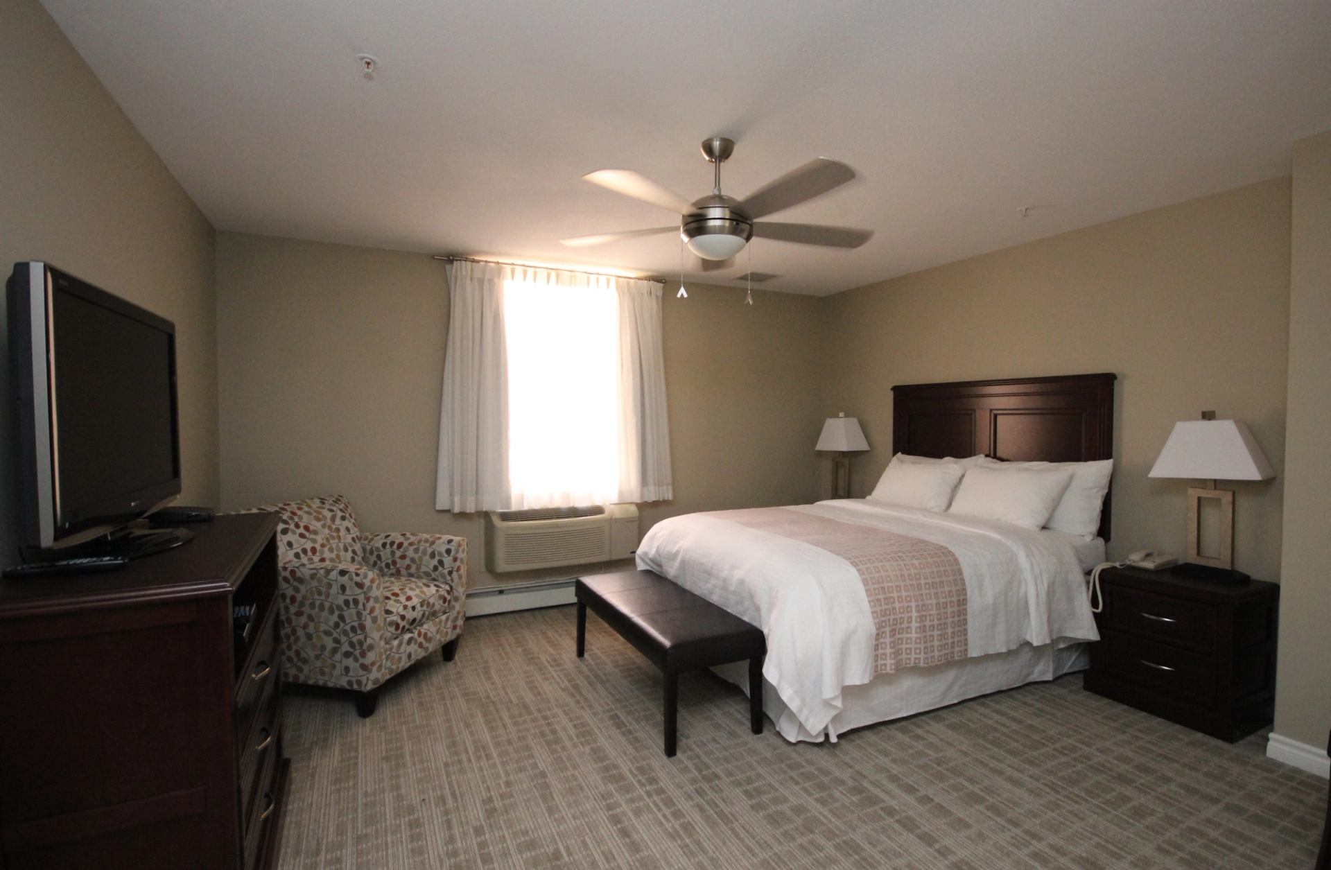 Hotel room with queen bed, TV, armchair, ceiling fan, lamps, and window with white curtains.