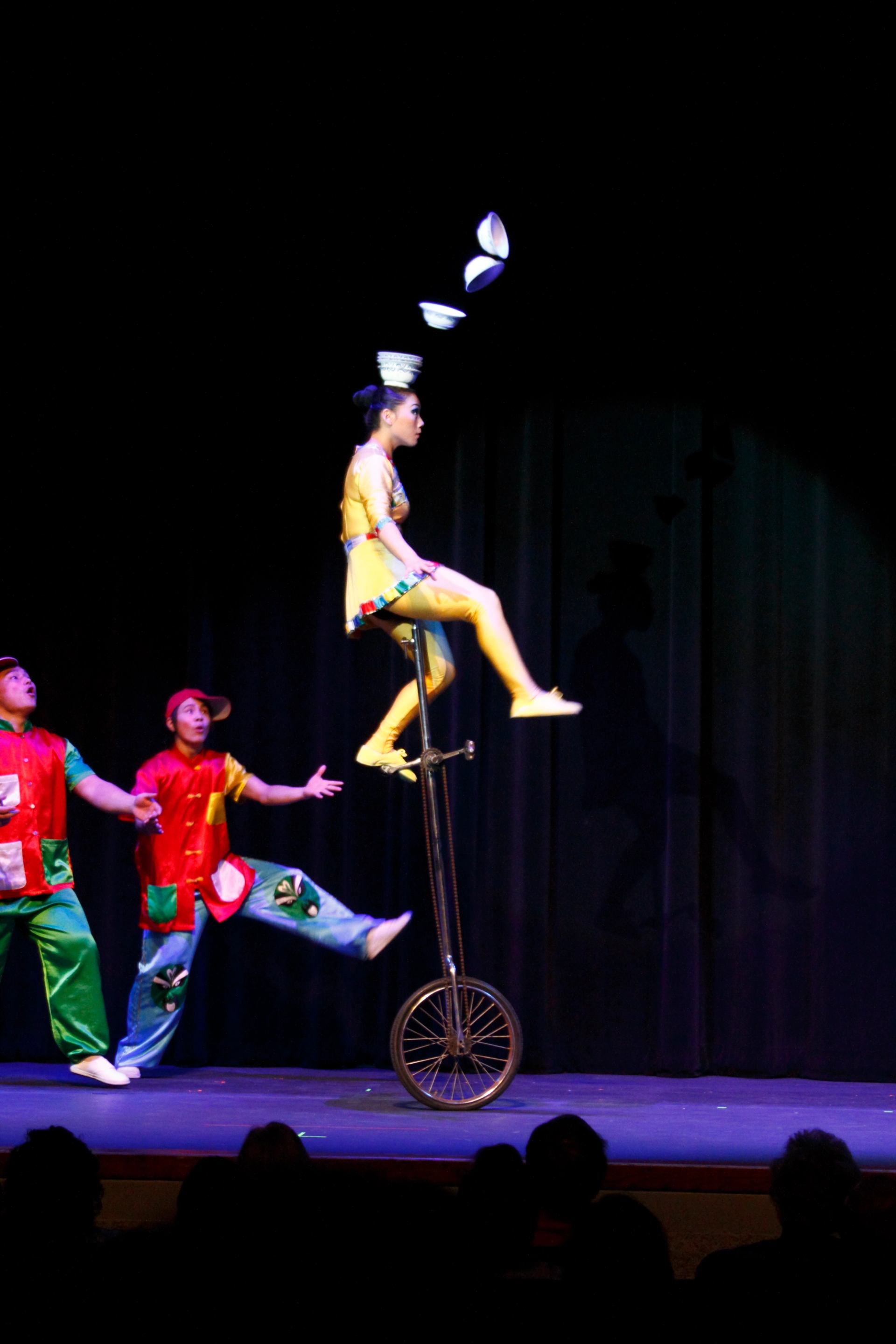 Circus performer rides tall unicycle while balancing and tossing bowls on stage.