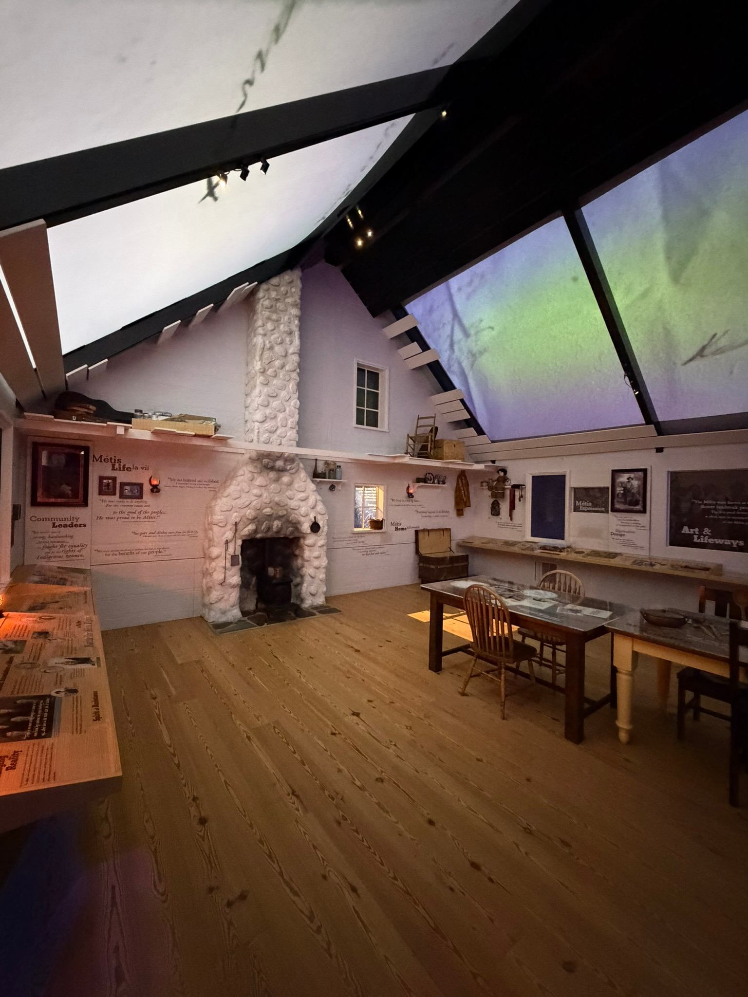Museum exhibit room with white stone fireplace, wooden tables, chairs and interpretive panels.