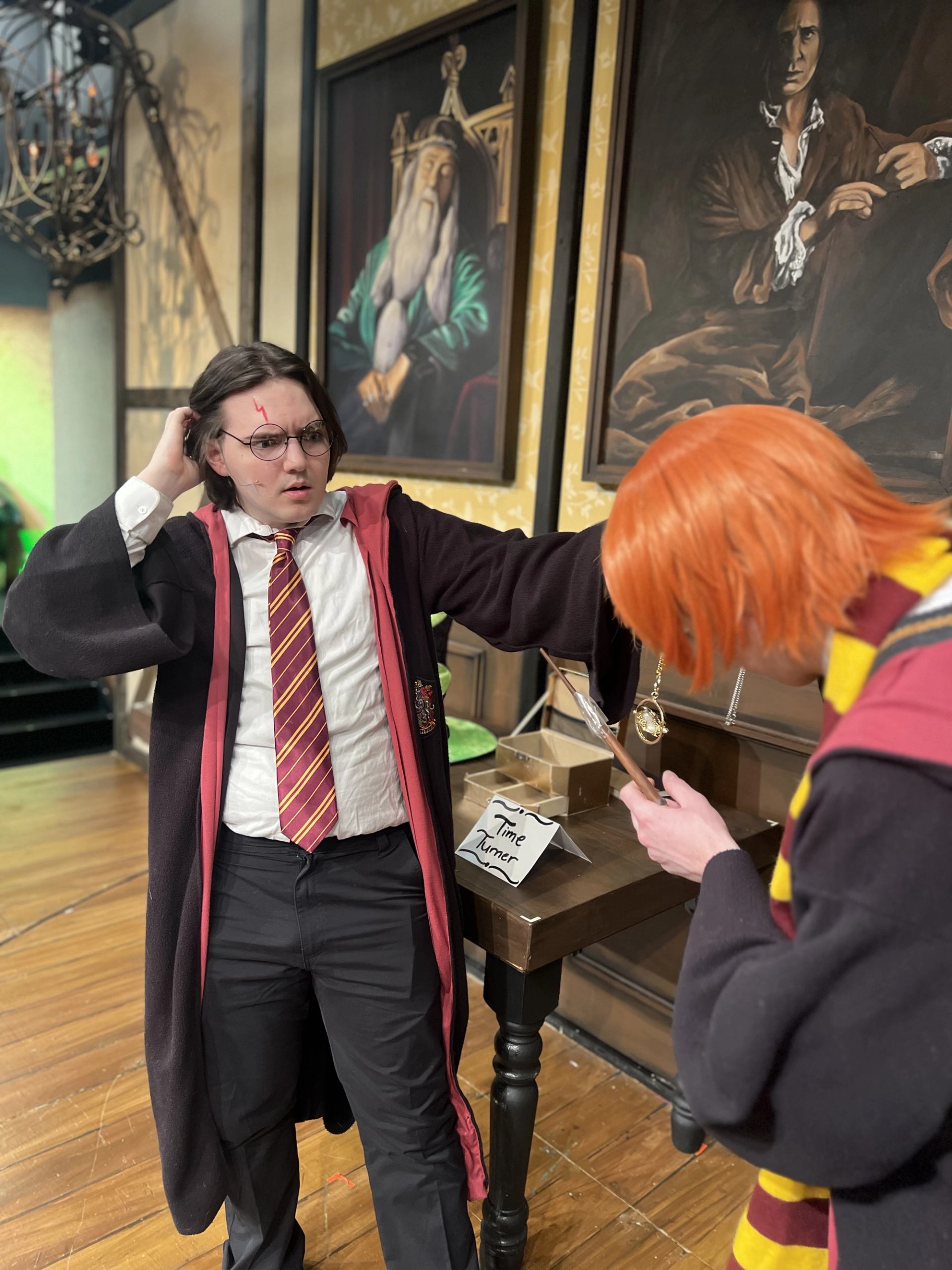 Two costumed people examine a Time Turner prop beside framed wizard portraits on a wall.