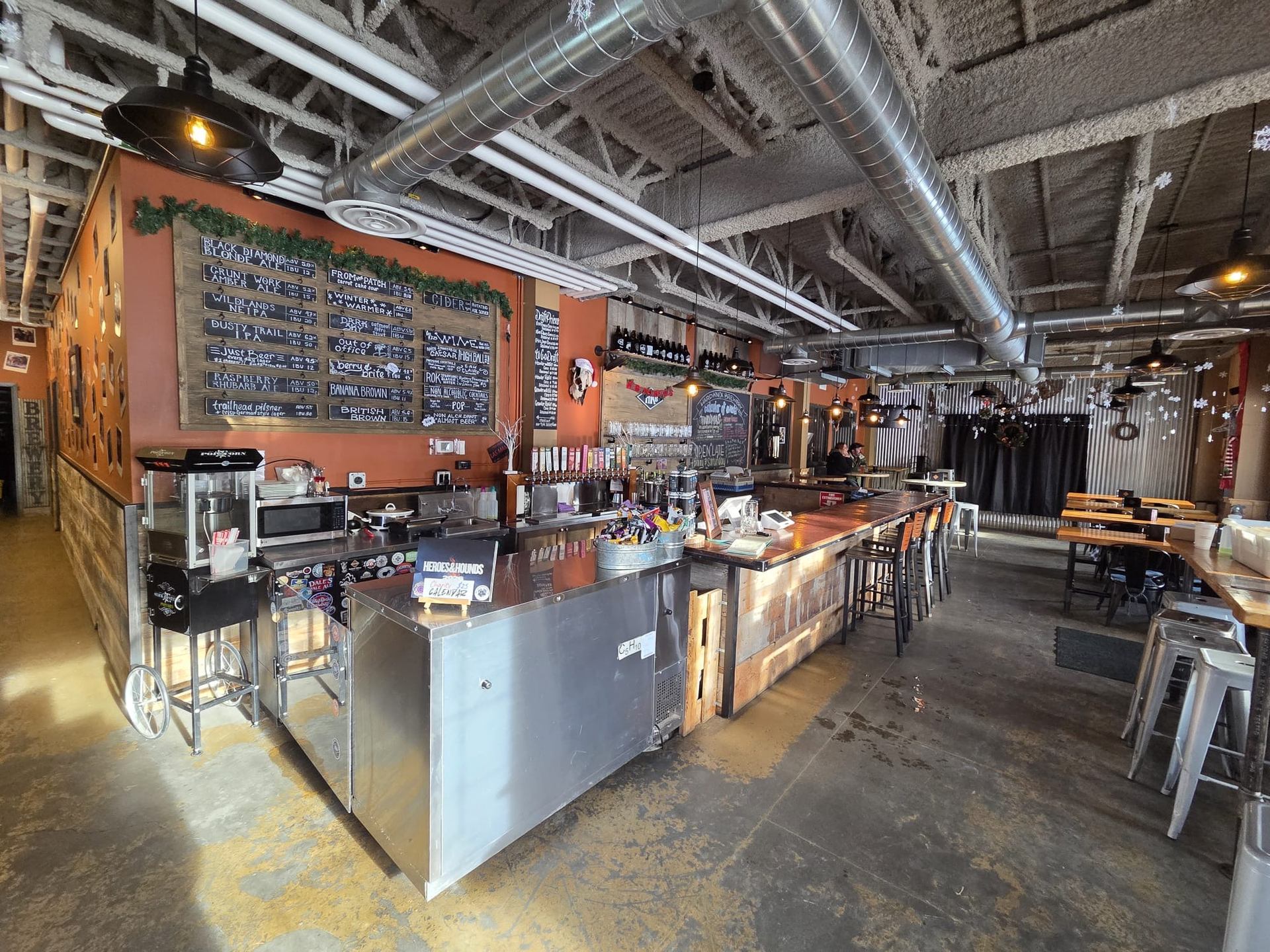The interior of a casual brewery or bar with a large chalkboard menu, a long bar, and exposed industrial ceilings.