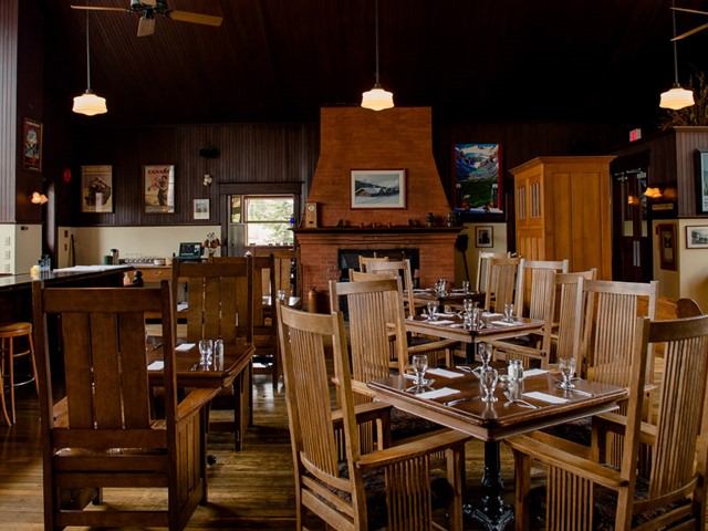 Cozy, dimly-lit restaurant interior with wooden tables and chairs. Warm tones, artwork on walls, and a central fireplace create an inviting atmosphere.