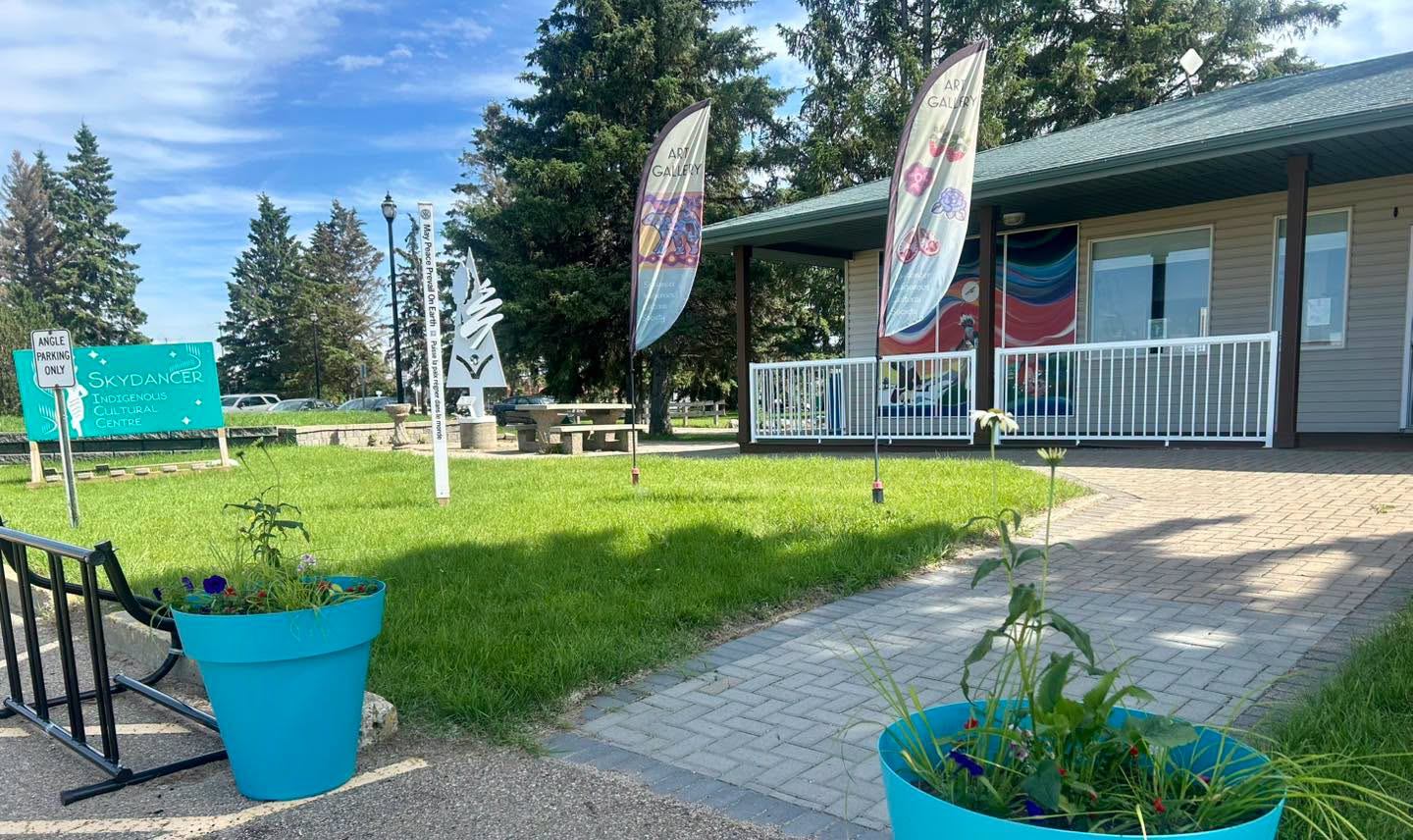 Skydancer Indigenous Cultural Centre exterior with art gallery flags, lawn, planters and trees.