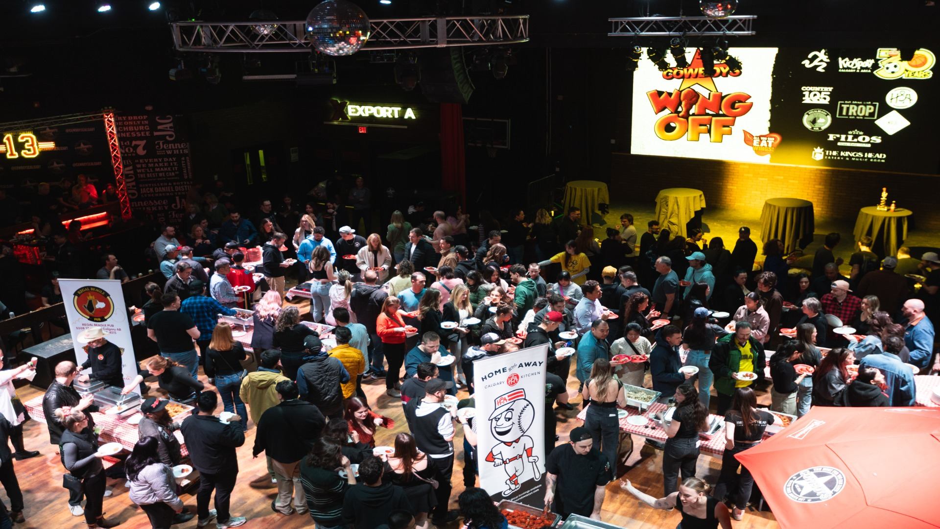 A large crowd fills the venue, moving between wing stations under bright event lighting.