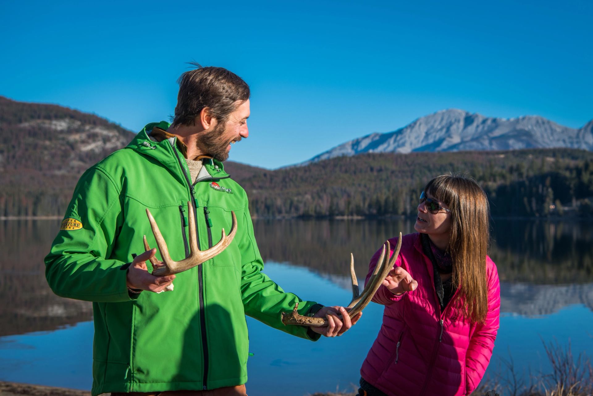 Two people holding deer antlers beside a calm lake with forested mountains.