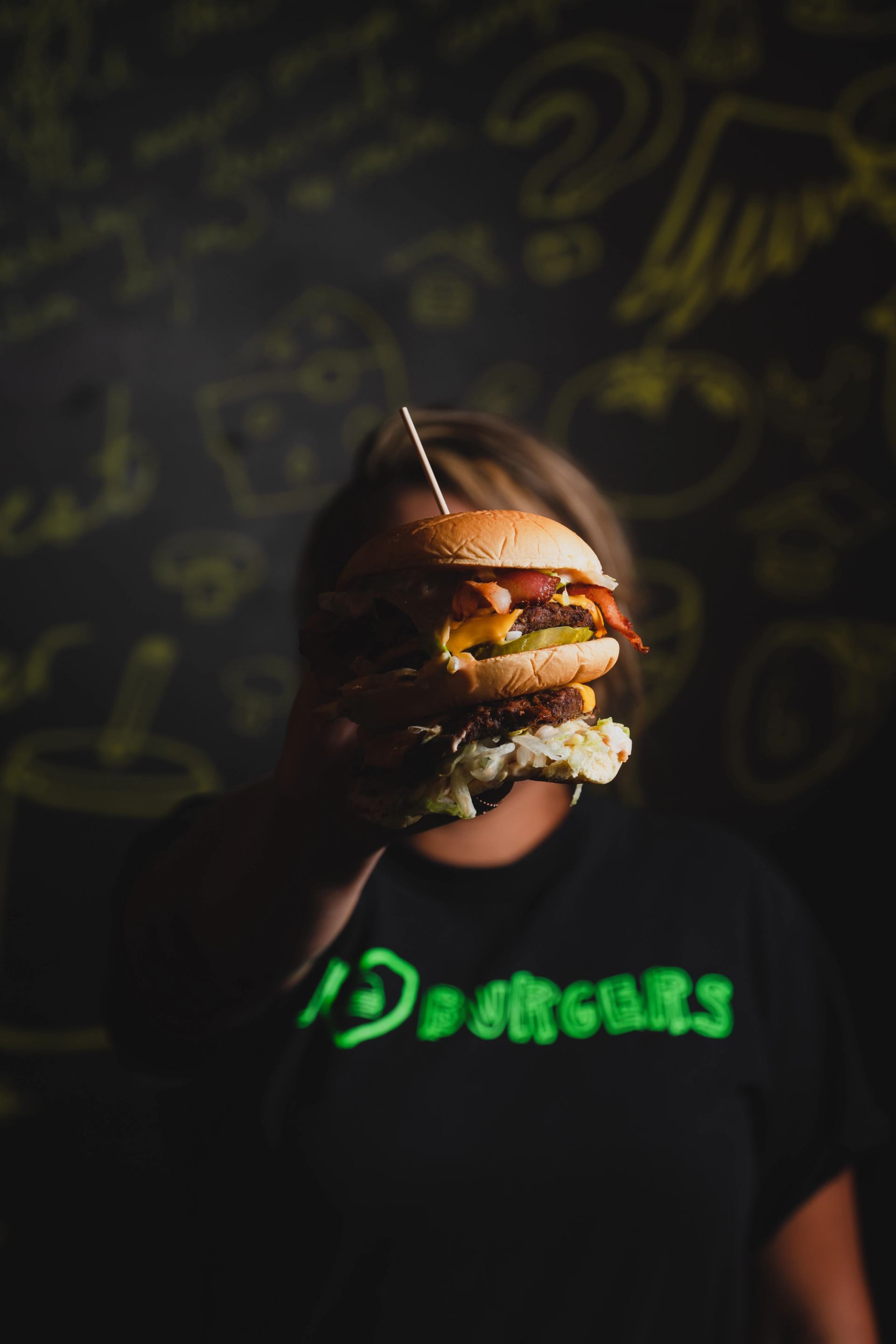 A person in a black "D BURGERS" shirt holds a large double bacon cheeseburger with pickles and lettuce in front of a dark, chalk-drawn wall.