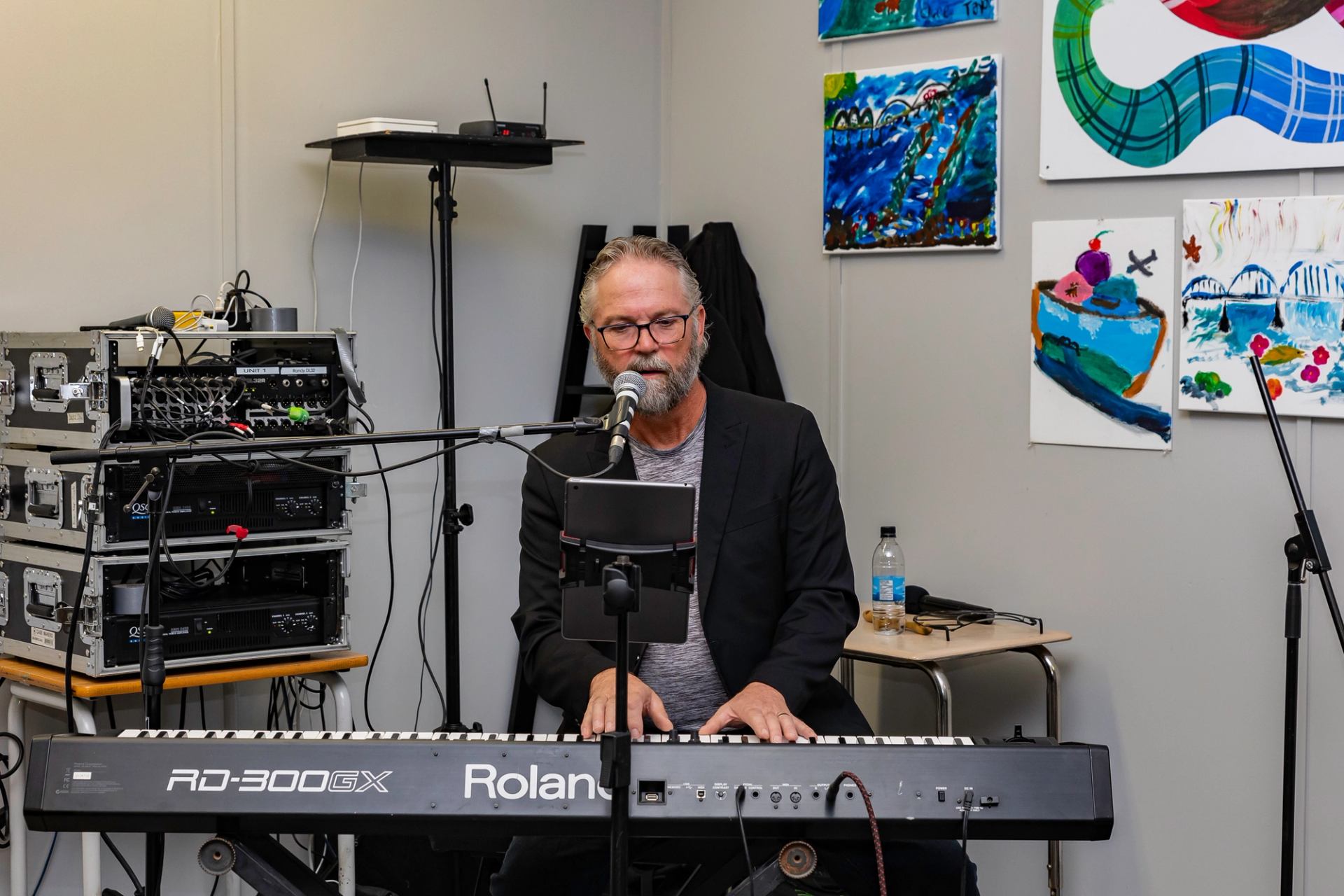 Man playing keyboard synthesizer at microphone in room with framed paintings on walls.