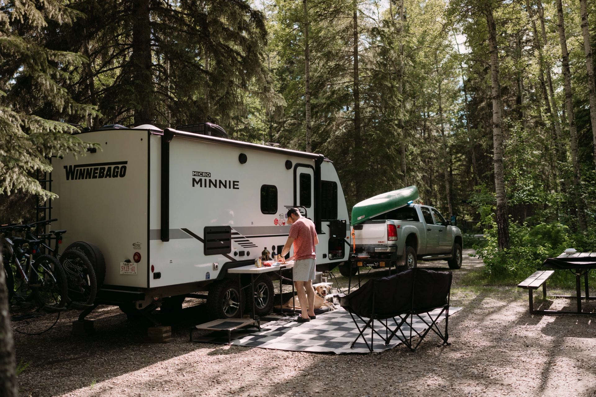 Campsite with Winnebago trailer, pickup truck carrying canoe, person cooking, dog resting.