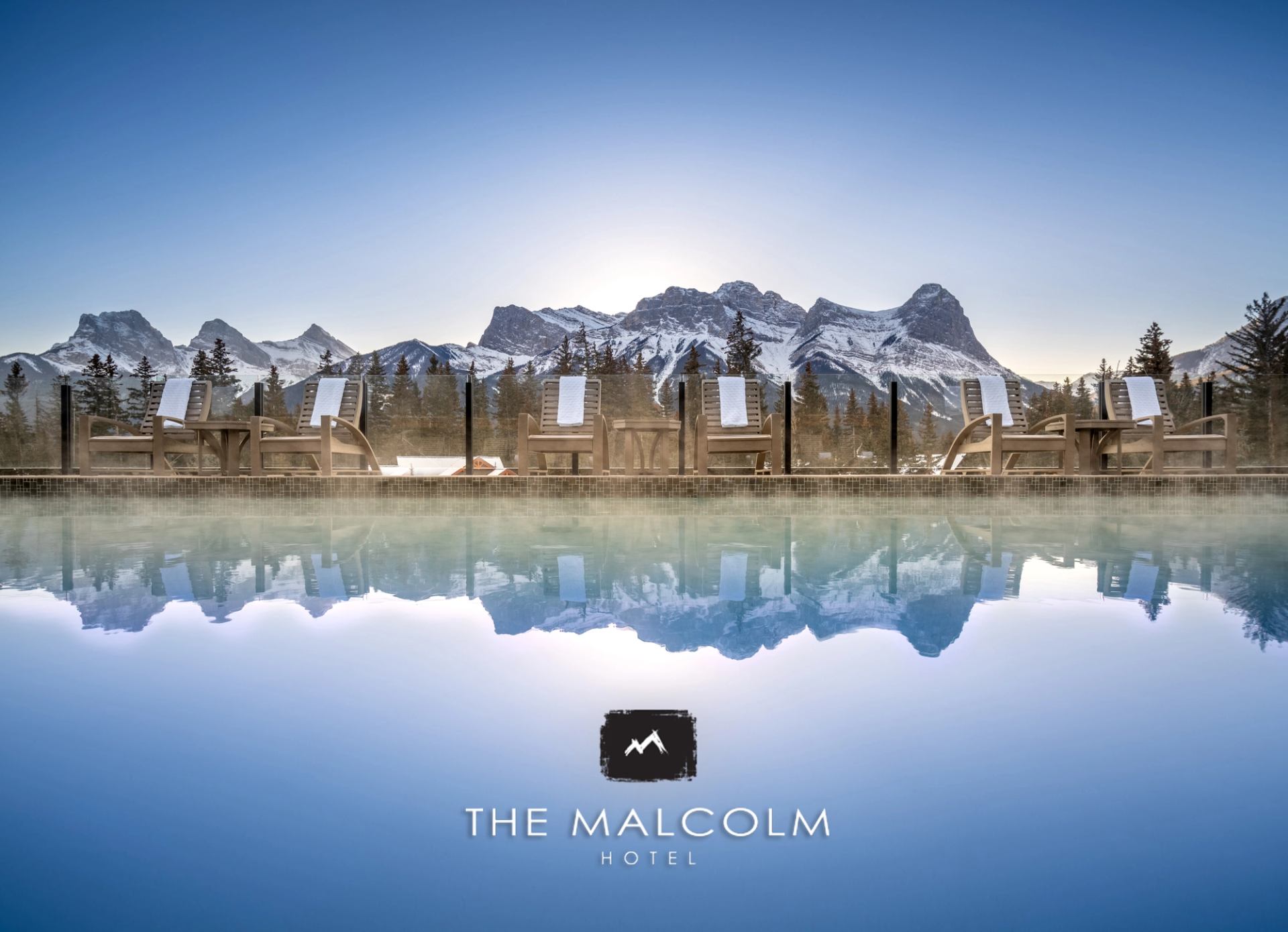Poolside chairs face snow-covered mountains reflected in water at The Malcolm Hotel.