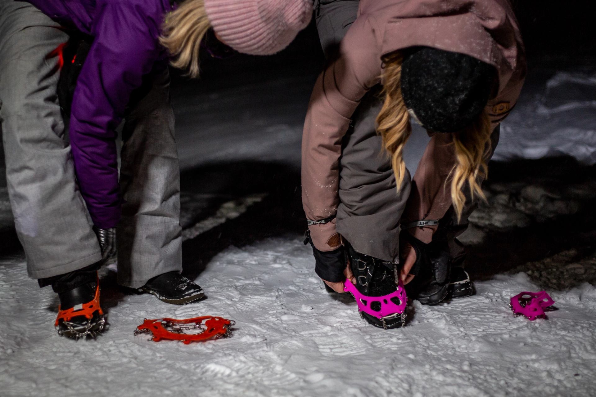 Two people fastening traction cleats onto boots on a snowy trail at night.