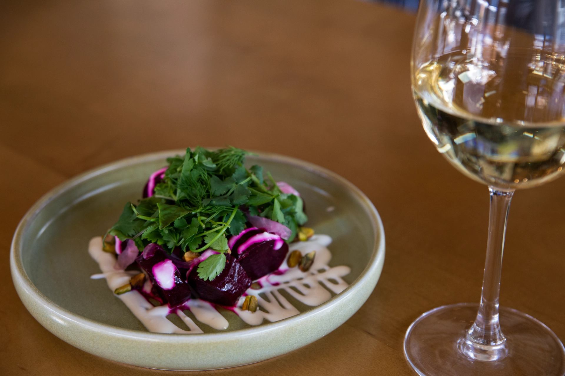 Plate of roasted beets with white sauce, pistachios, and herbs, next to a glass of white wine.