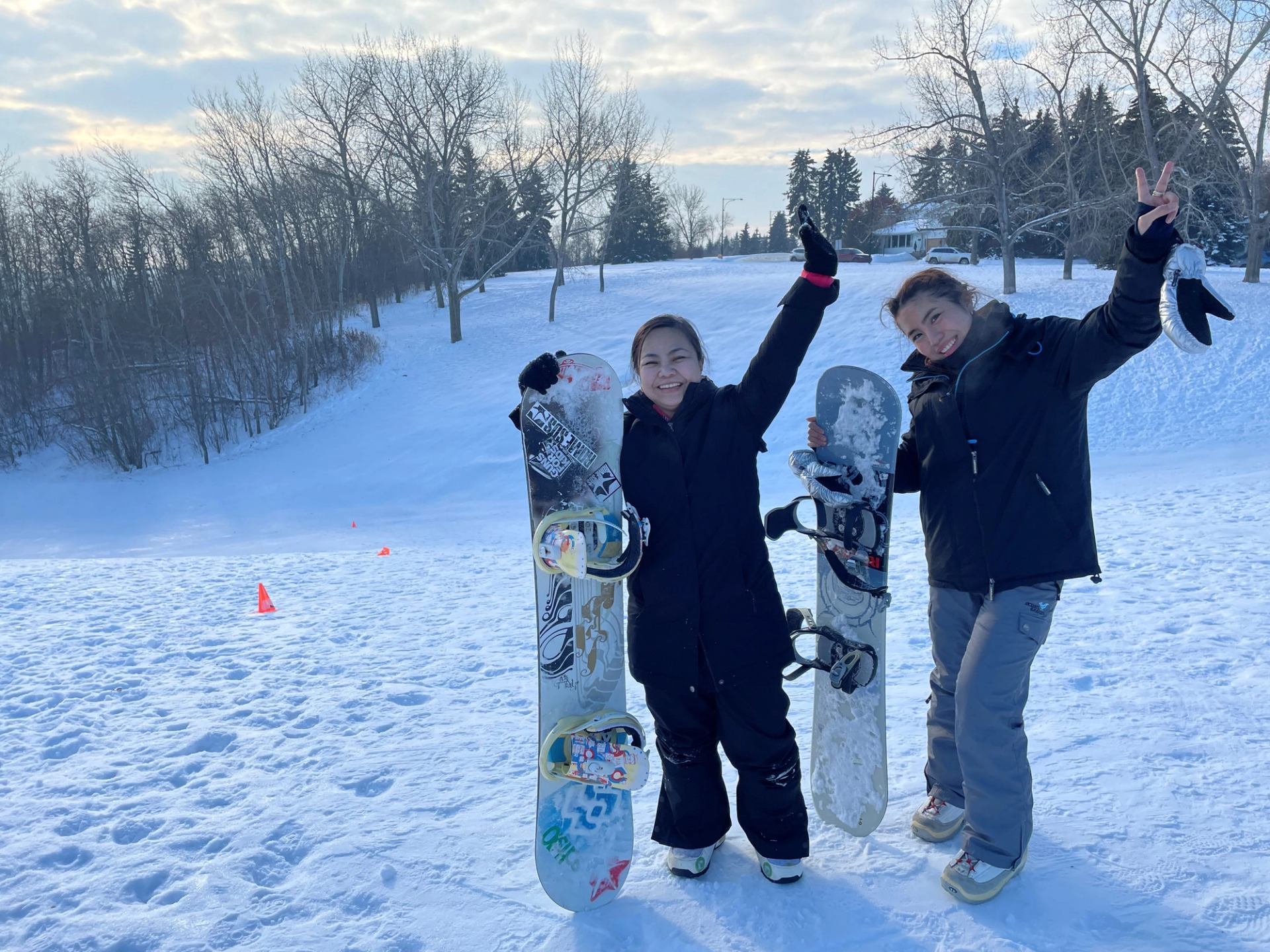 Snowboarders pose with snowboards on a snowy field with orange cones and bare trees behind.