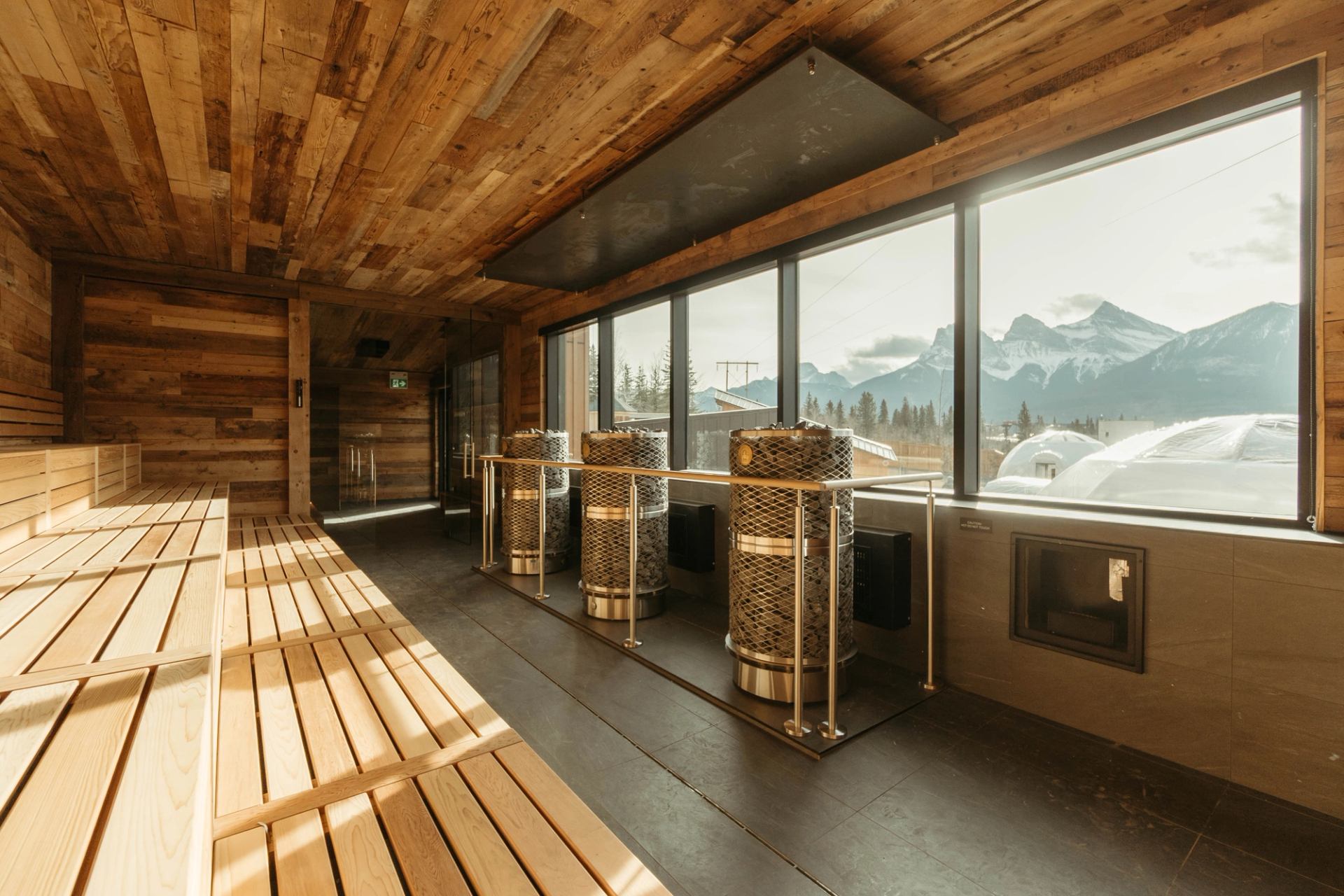 Wood-panelled sauna interior with benches, metal heaters and large windows overlooking snowy mountains.