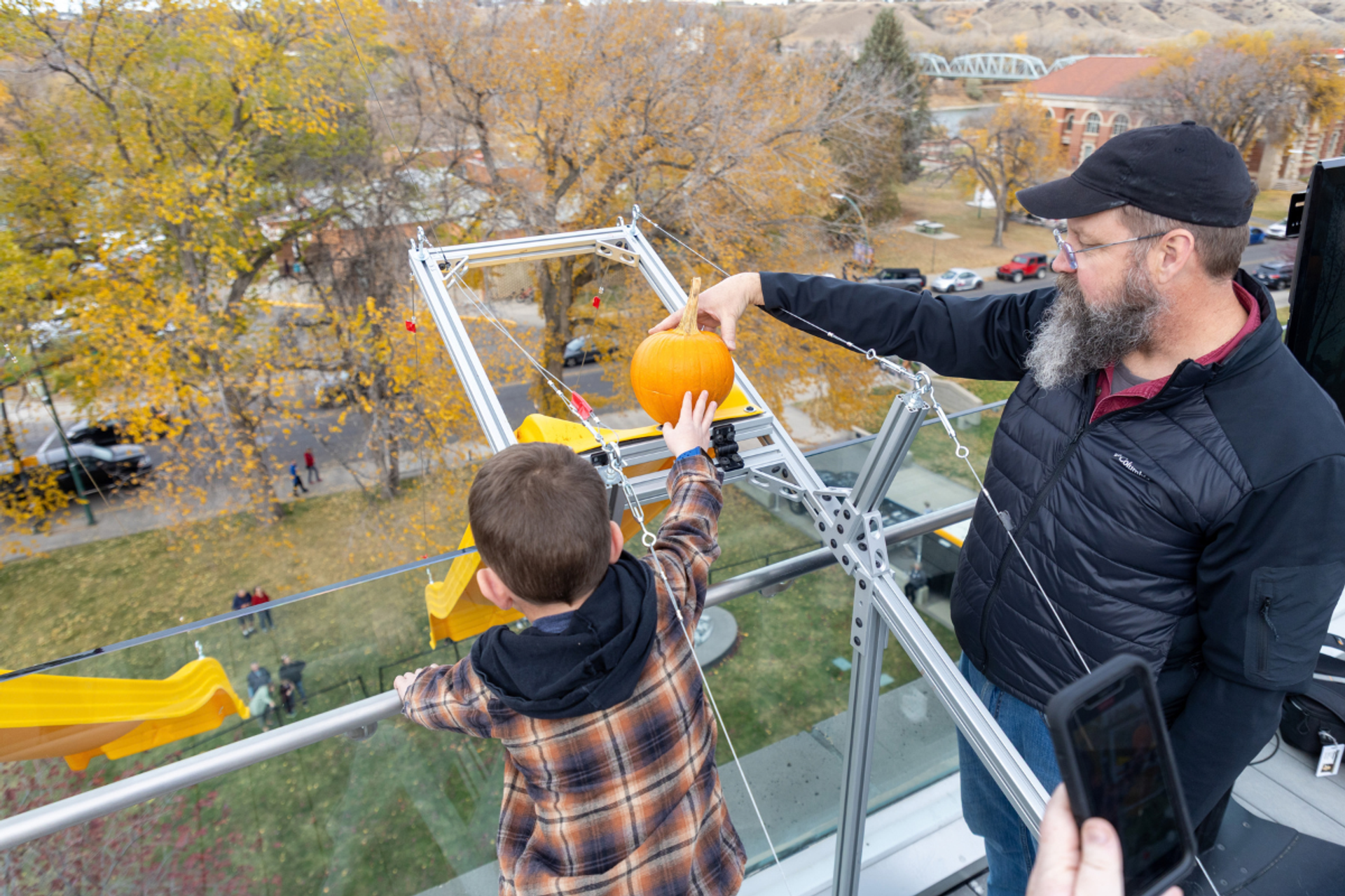 Man and child place a pumpkin on a metal frame above a glass railing.