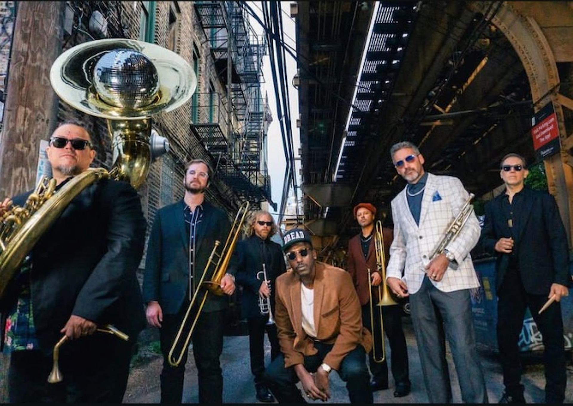 Brass band poses in urban alley under elevated tracks with instruments and disco-ball tuba.