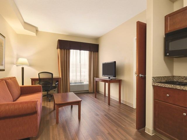 Hotel suite living area with sofa, desk, TV, kitchenette, and window curtains.