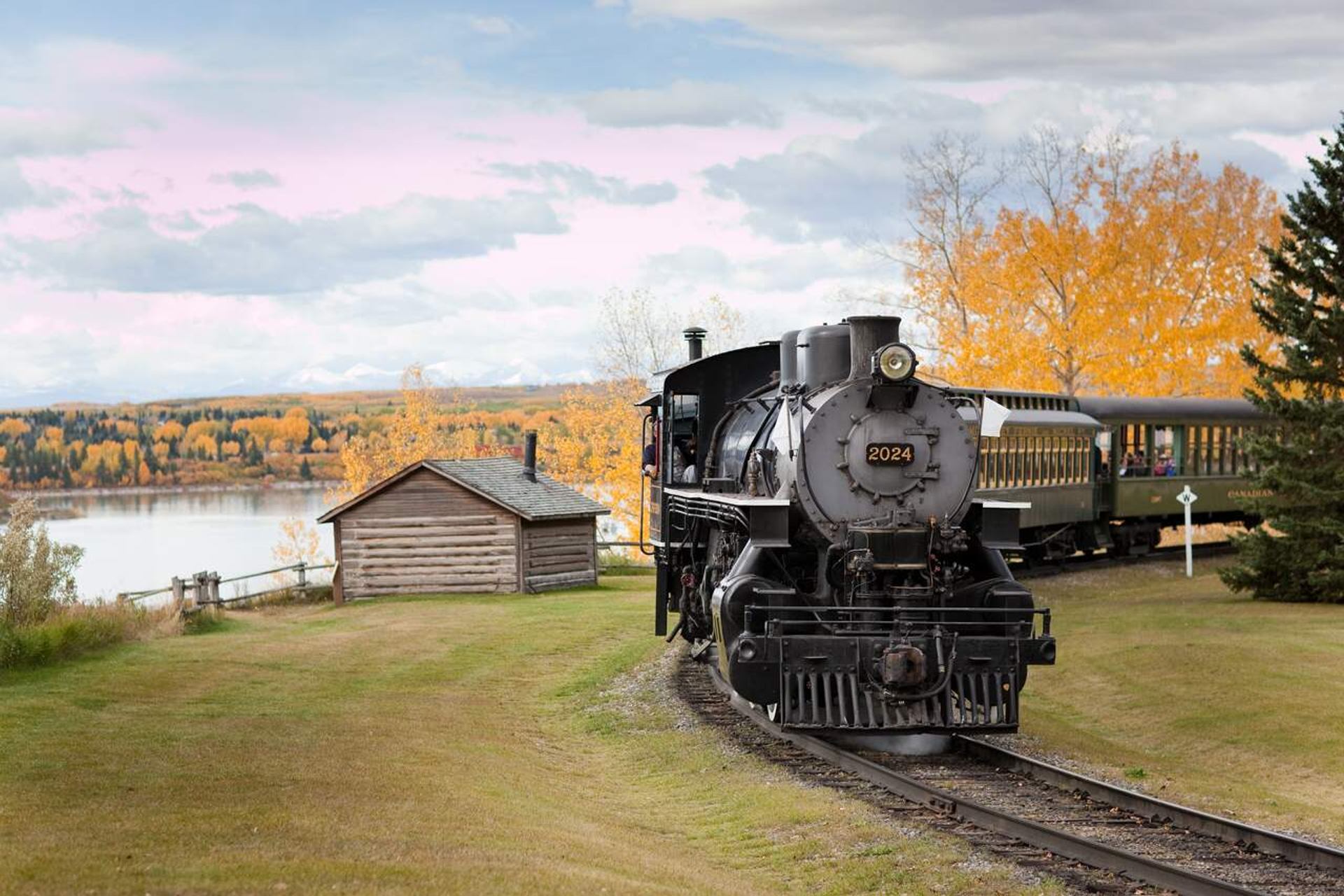 Steam locomotive 2024 passes a small log cabin beside a lake and trees.