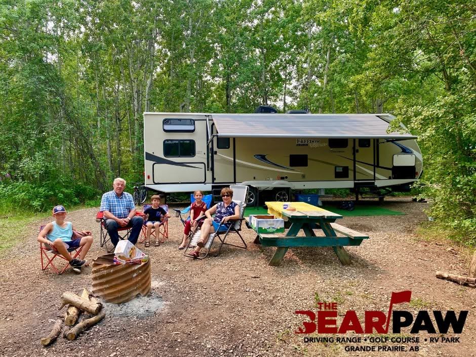 Family sits in camp chairs by firepit near RV and picnic table at Bear Paw.