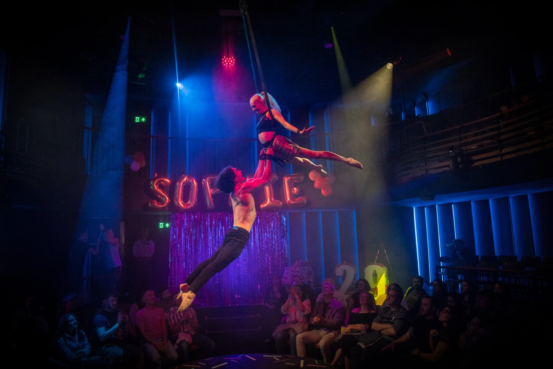Aerial performers hang above seated audience in colourful stage lighting and party decorations.