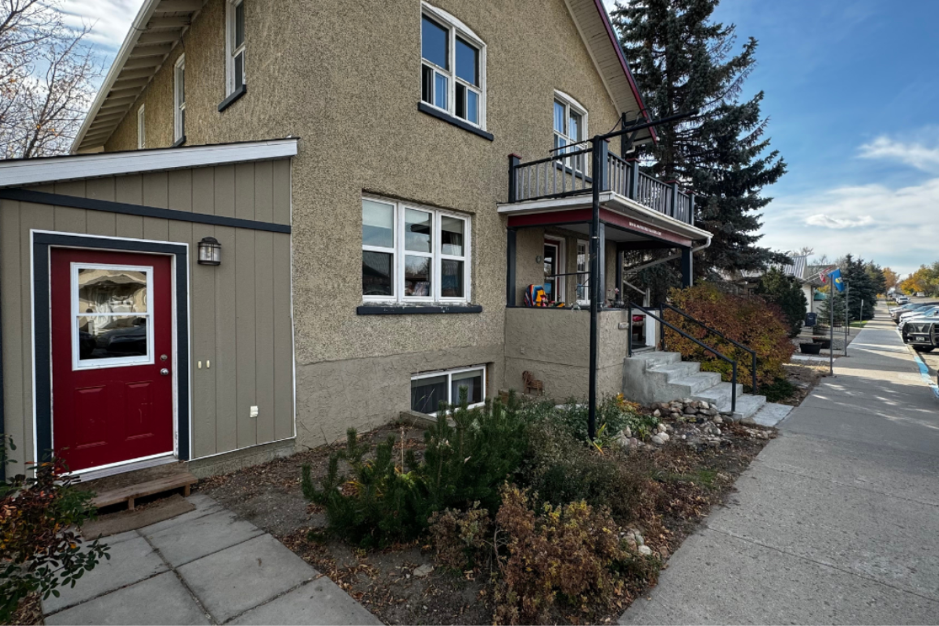 Two-storey stucco house with red side door, front steps, shrubs and sidewalk.