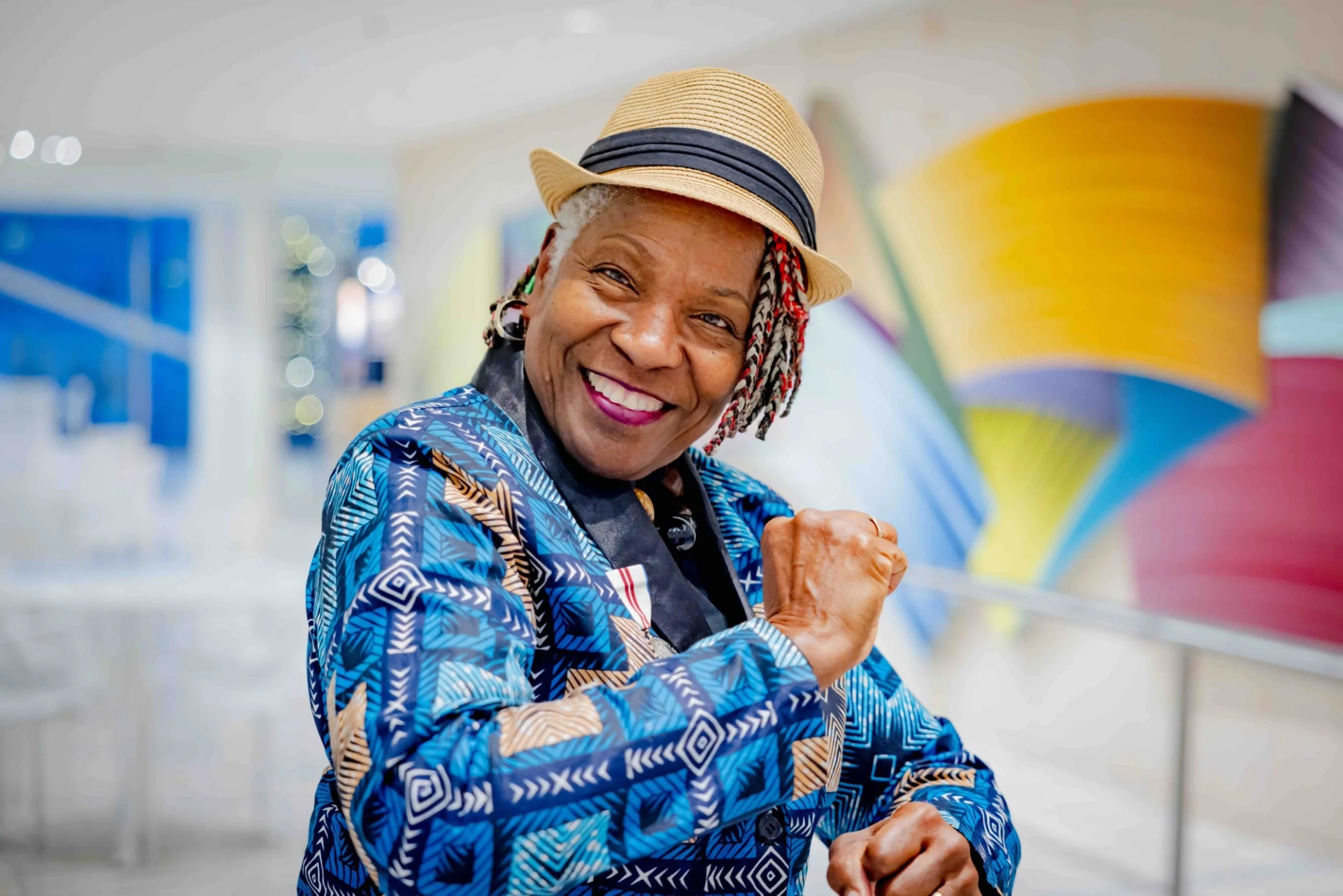 A smiling woman in a straw hat and blue patterned jacket raises her fist.