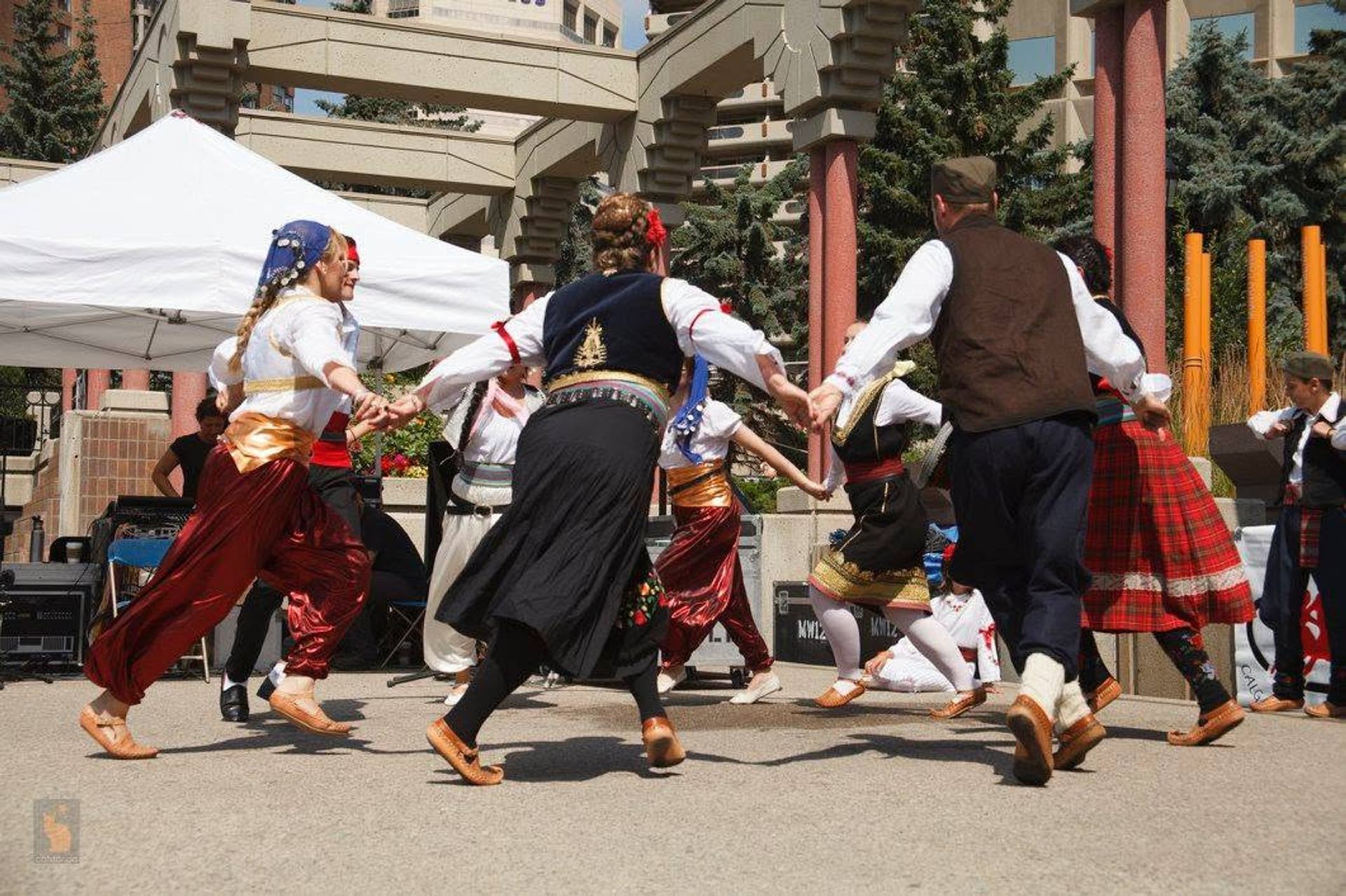Folk dancers in traditional costumes hold hands in a circle at an outdoor event.