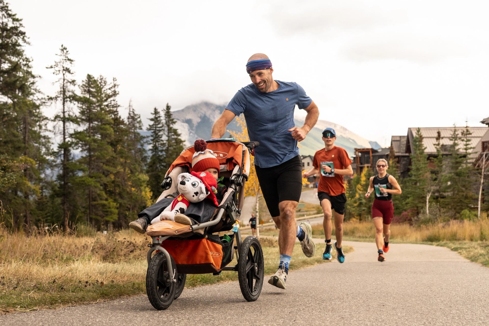 Runner pushes stroller with child and stuffed dog on paved path near forested mountains.