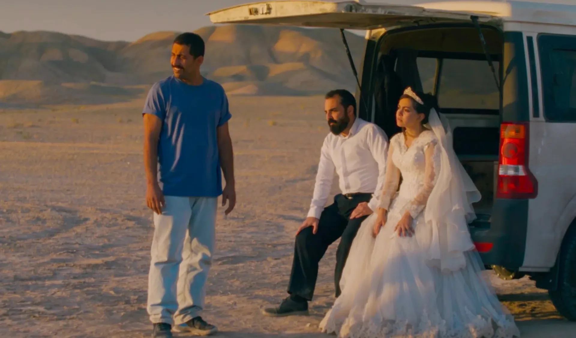 A bride and groom sit at the back of an open SUV in a desert landscape while another person stands nearby.