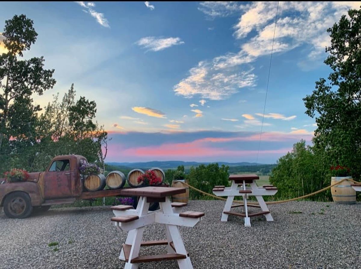 Gravel patio with picnic tables, old truck, barrels and distant hills under colourful sky.