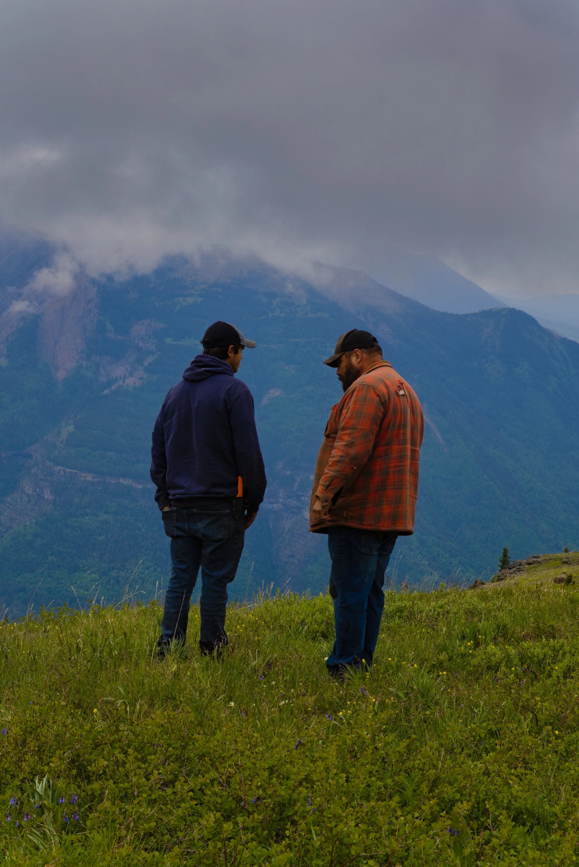 Two people standing on a grassy hill with mountains and clouds in the background.