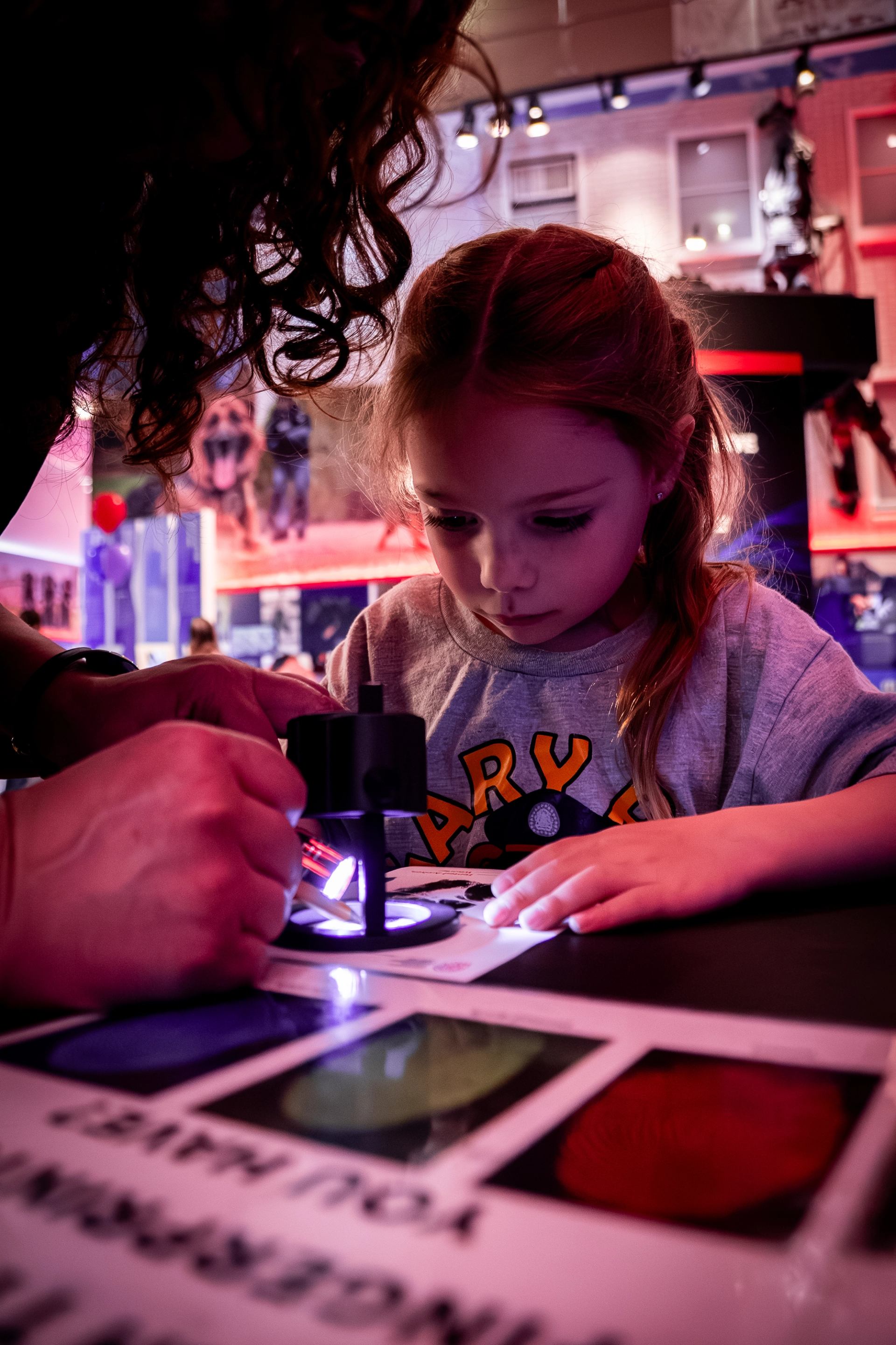 Child examines paper under small microscope with adult assistance in colourful indoor exhibit.