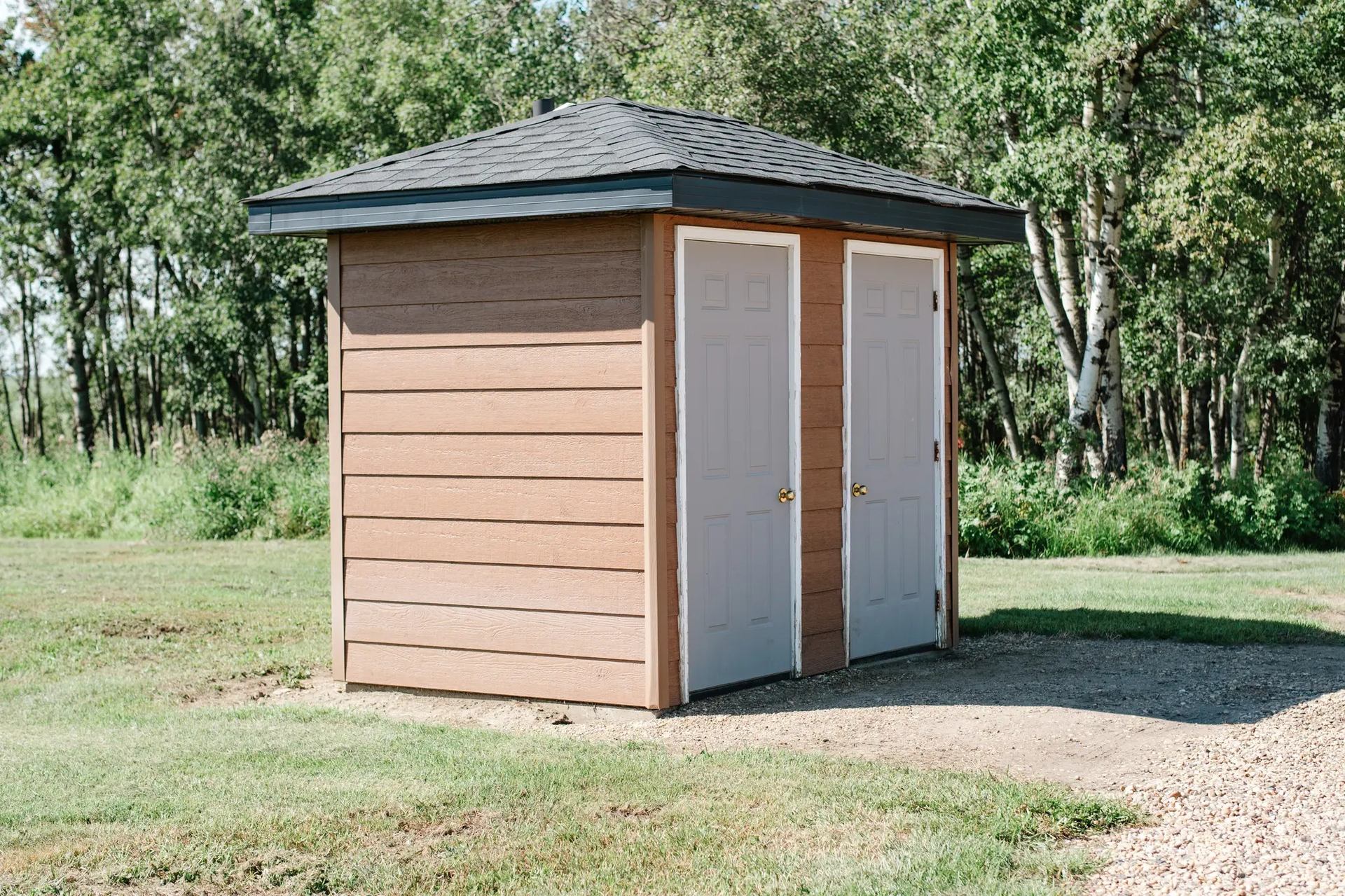 Small brown outbuilding with two white doors, standing on grass near trees.