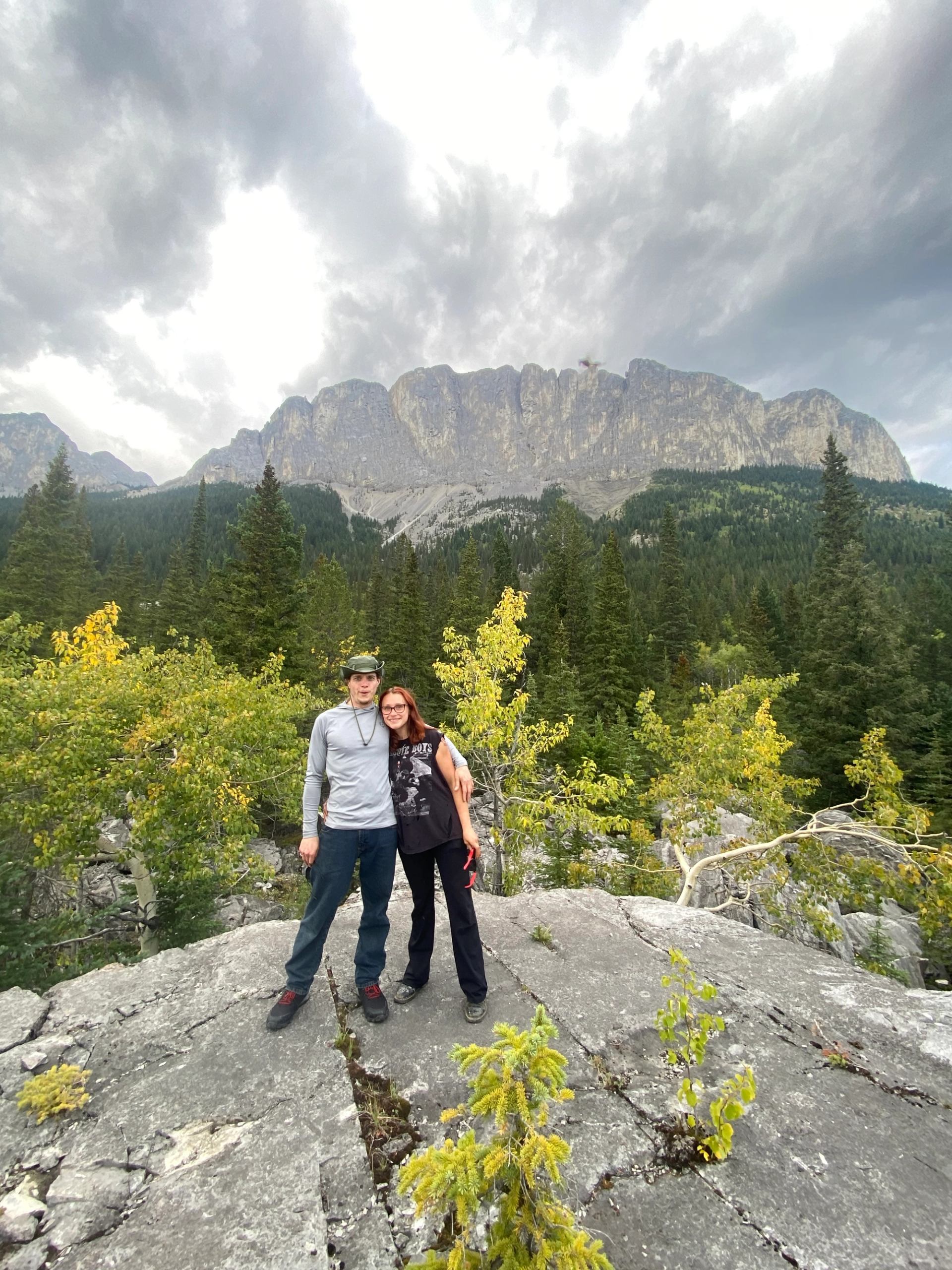 Two hikers stand on rocky outcrop above dense forest with towering cliffs behind.