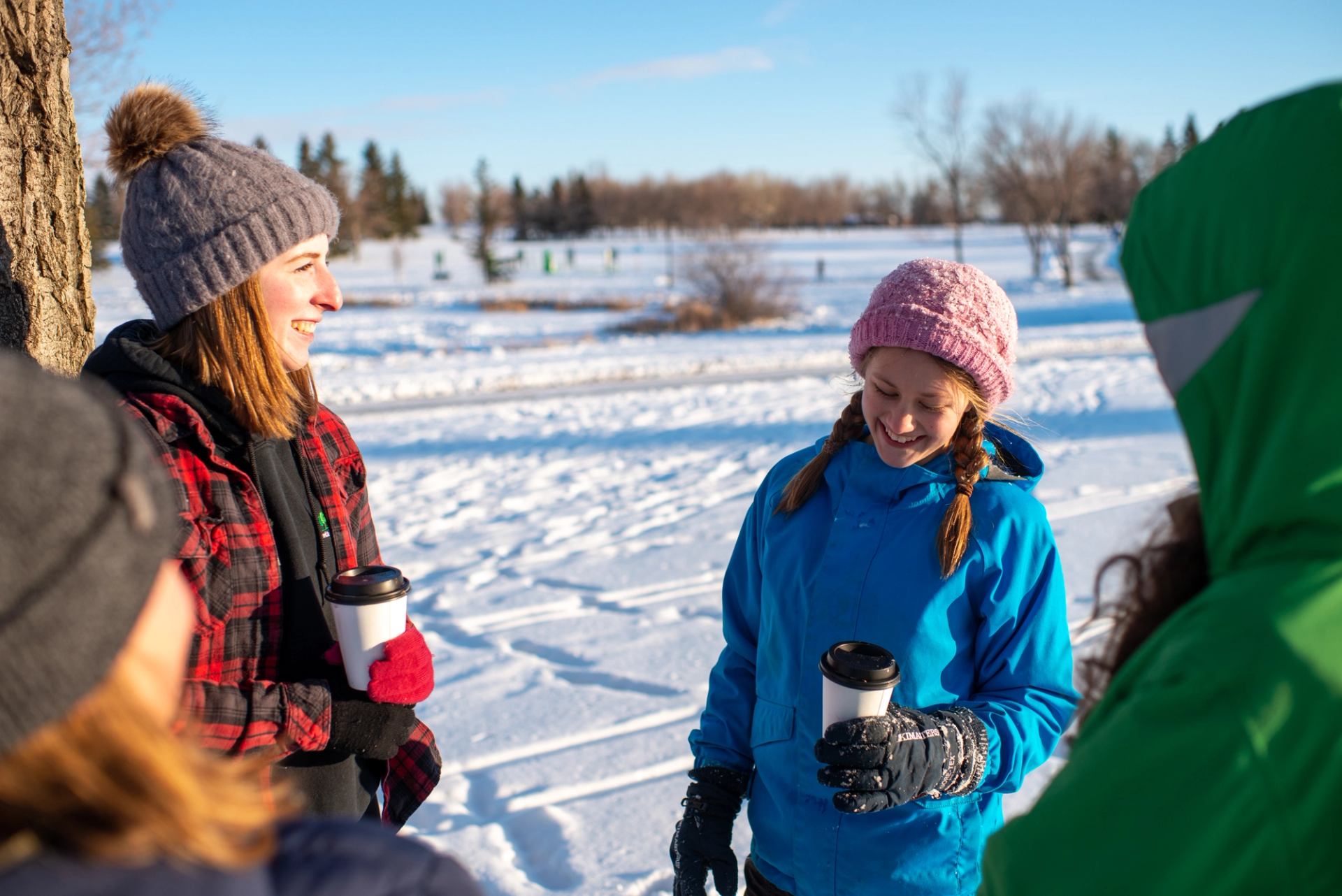 Three smiling people holding coffee cups in a sunny, snowy park.