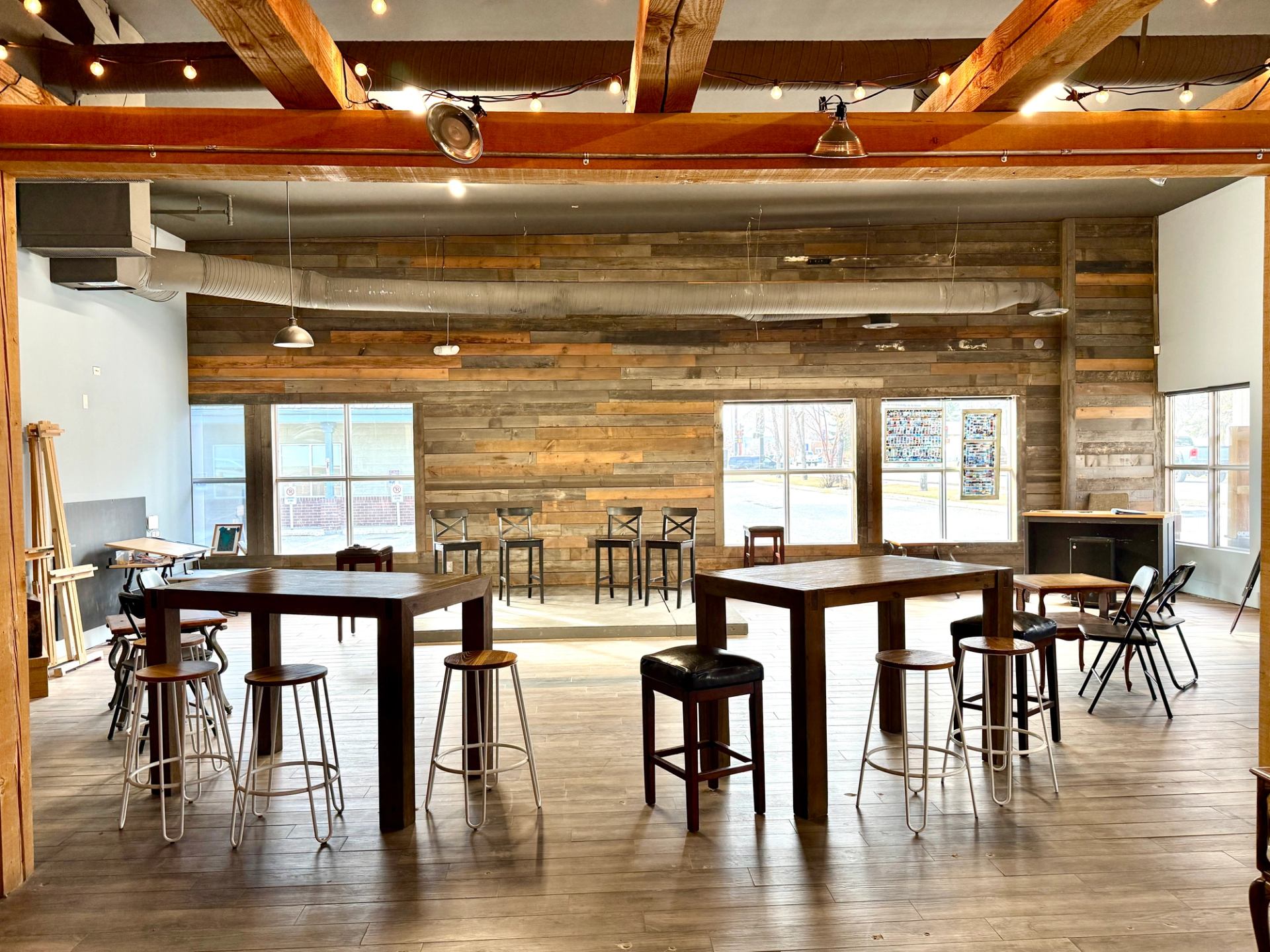 Spacious room with wooden tables, stools, folding chairs and string lights overhead.