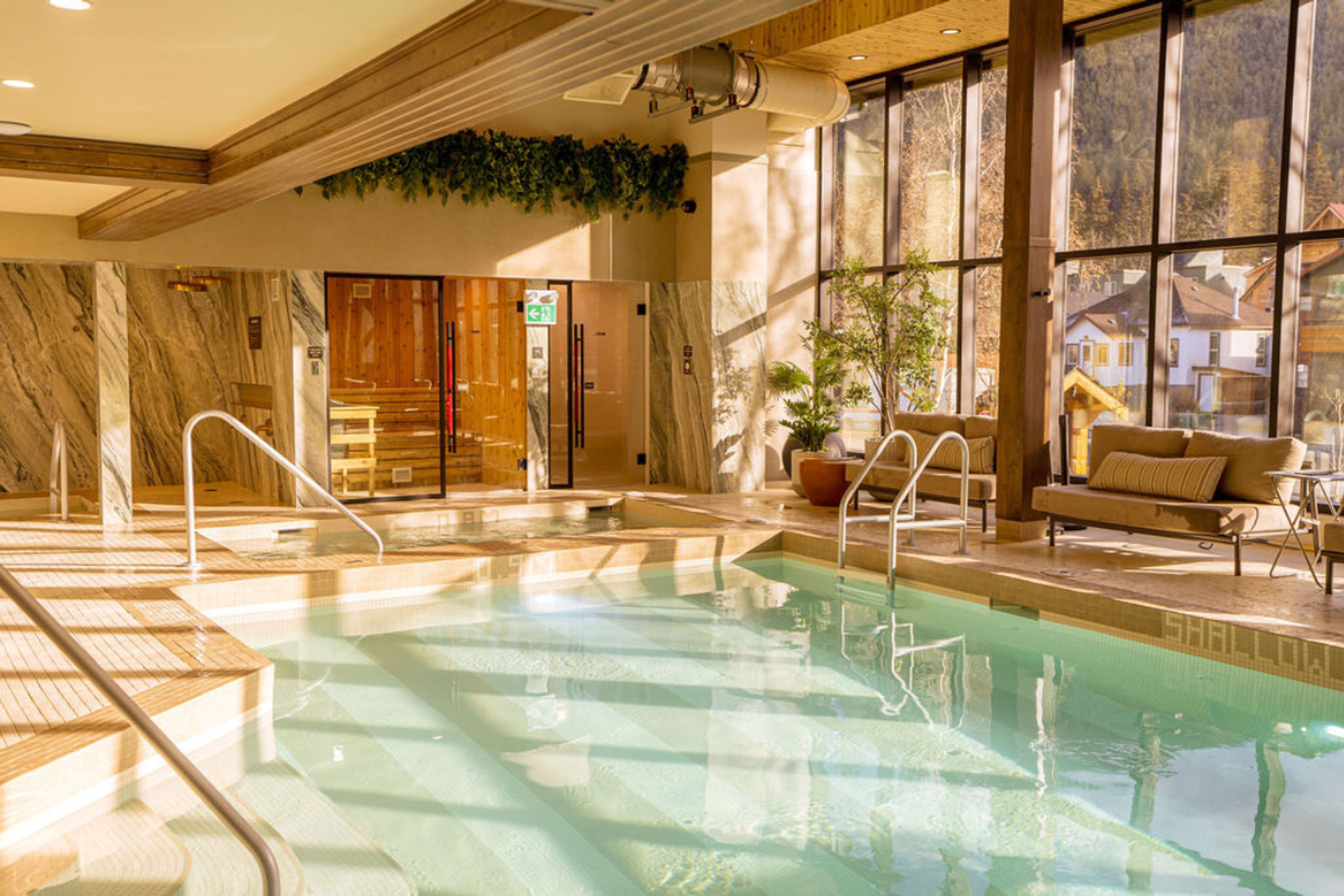 Indoor pool area with a hot tub, sauna, and lounge seating next to large windows overlooking a village.