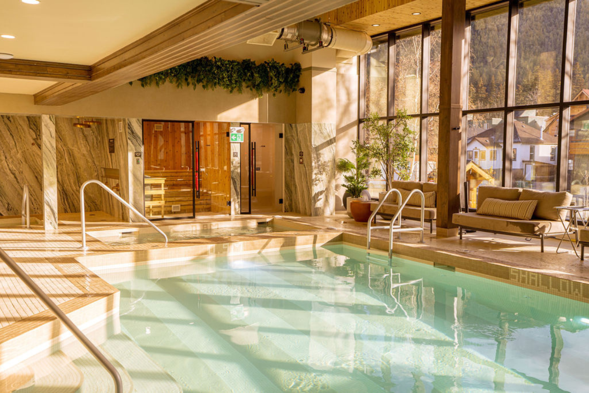Indoor pool area with a hot tub, sauna, and lounge seating next to large windows overlooking a village.