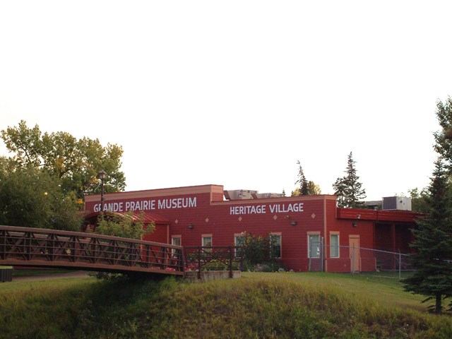 Red museum building with bridge and greenery at Grande Prairie Heritage Village.