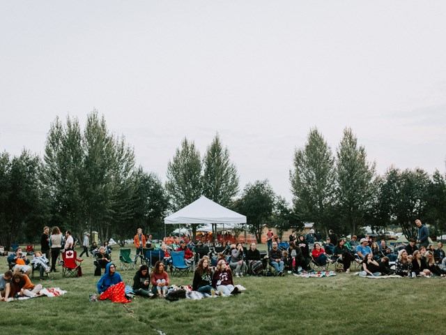 Backyard festival crowd seated on grass near white event tent and trees.